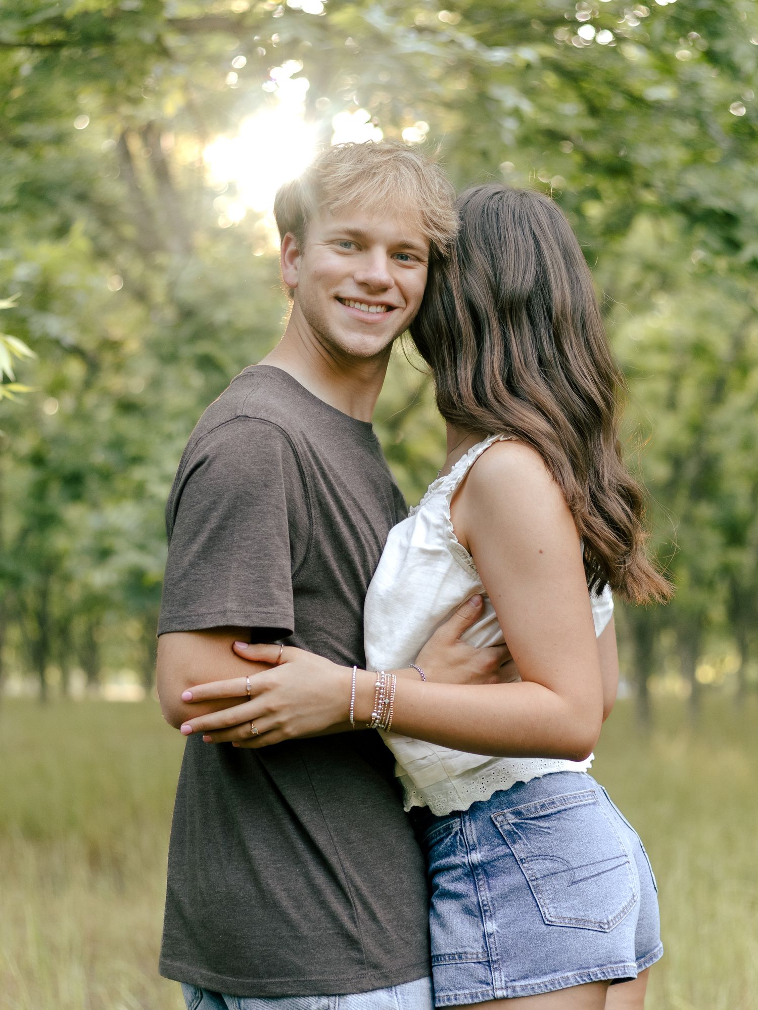 Smiling young man in a gray t-shirt embraces a woman in a denim skirt and white top in a sunlit grassy field with trees.