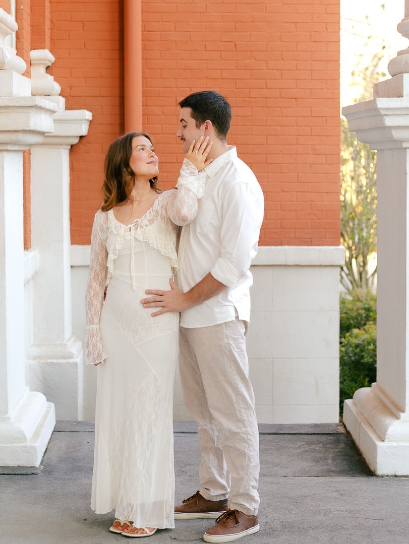 Couple embracing in front of a red brick building with white columns, she wears a lace white dress and he wears a white shirt and khakis.