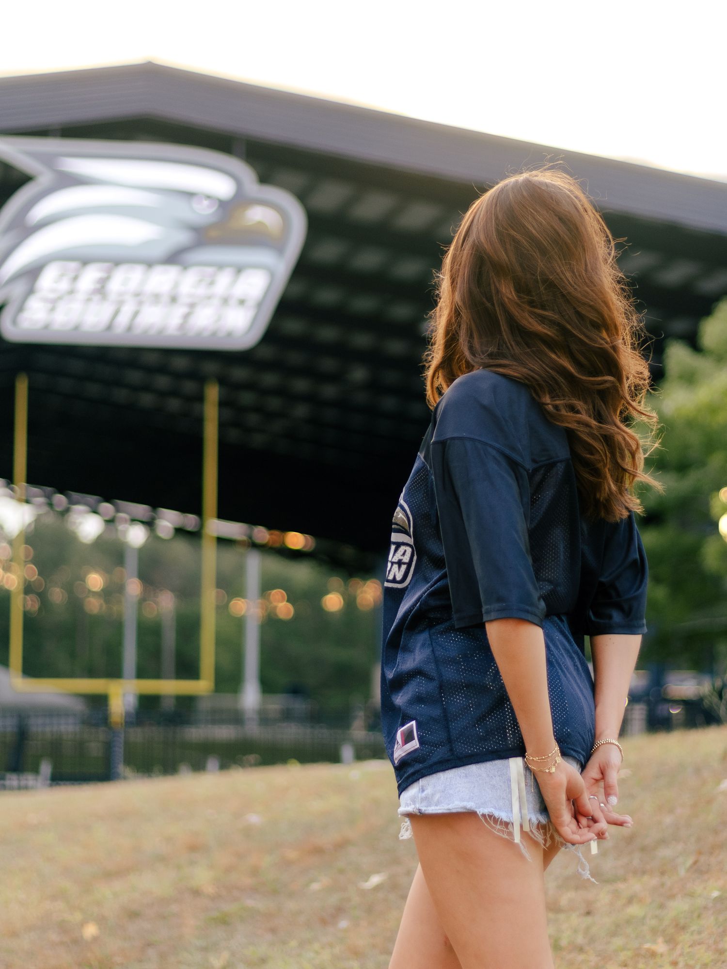 Woman in a navy football jersey and denim shorts stands facing a Georgia Southern stadium on a grassy hill at sunset.
