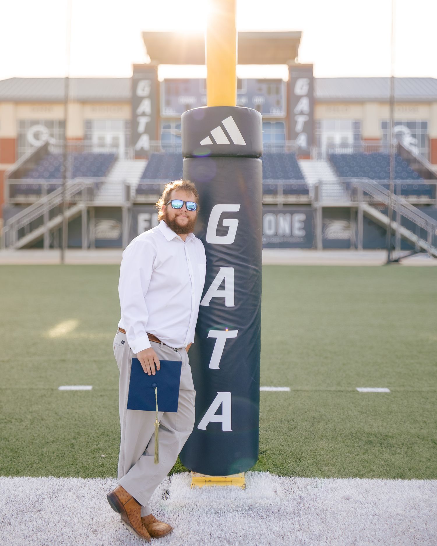 Man in sunglasses, white shirt and khakis leans on a padded goalpost marked GATA while holding a navy folder on a football field.