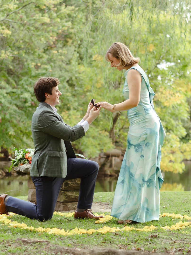 A man in a green blazer kneels on grass lined with yellow petals, holding a ring box as a smiling woman in a teal dress looks on.