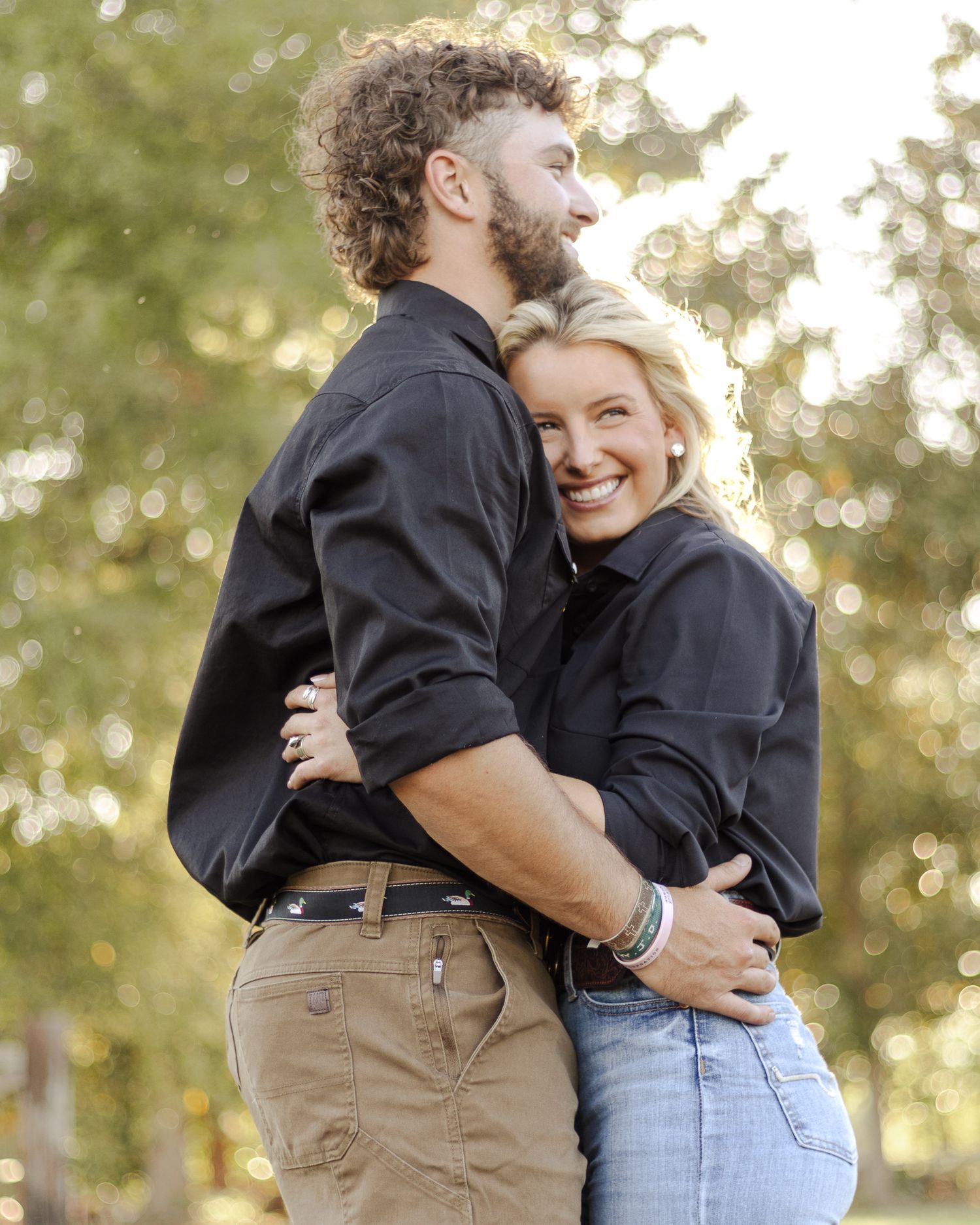 A smiling couple in matching dark button-up shirts embraces outdoors, with golden sunlight filtering through green trees behind them.