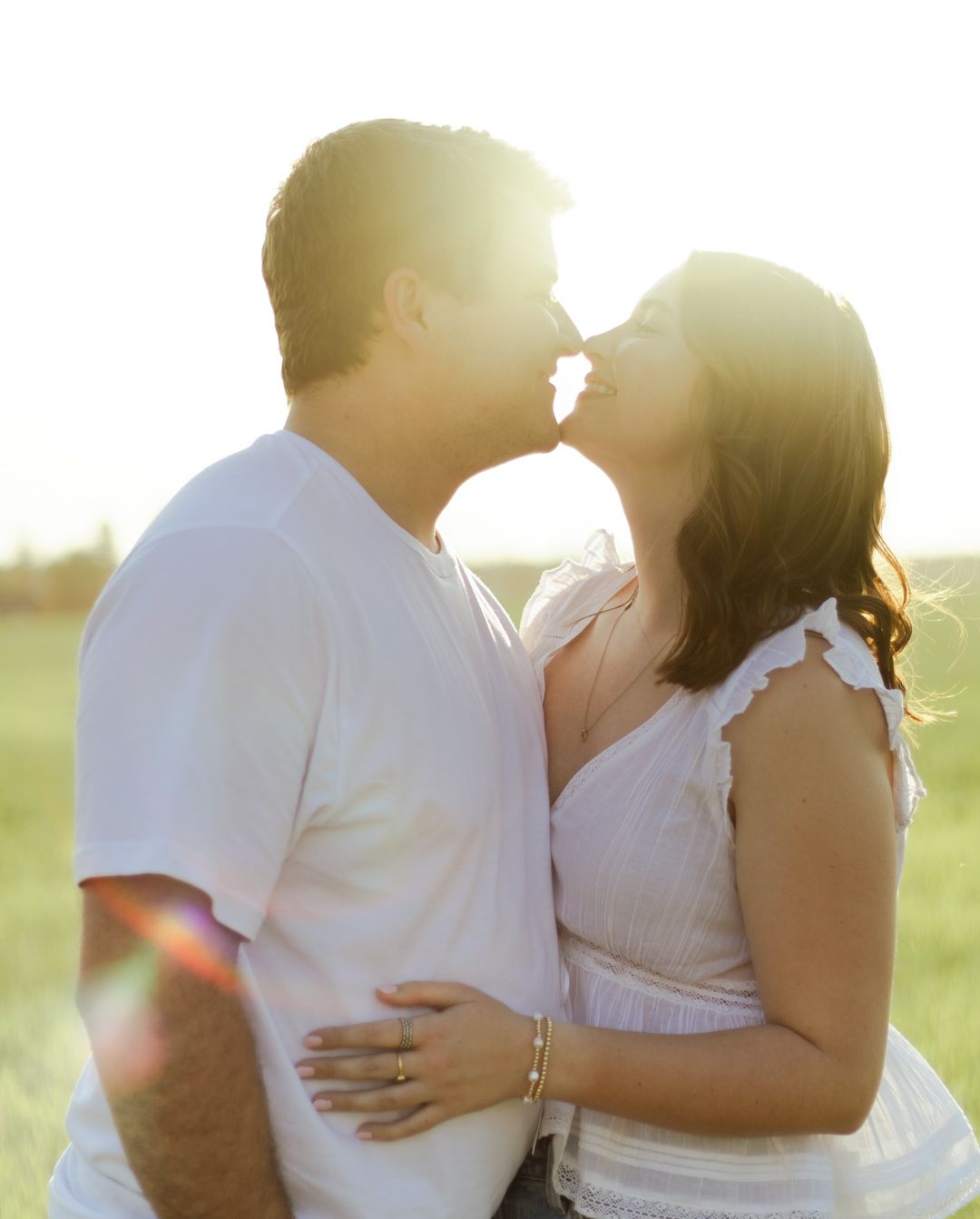 Couple sharing a romantic nose-to-nose moment in a sunlit green field, the woman in a white ruffled dress and the man in a white t-shirt.