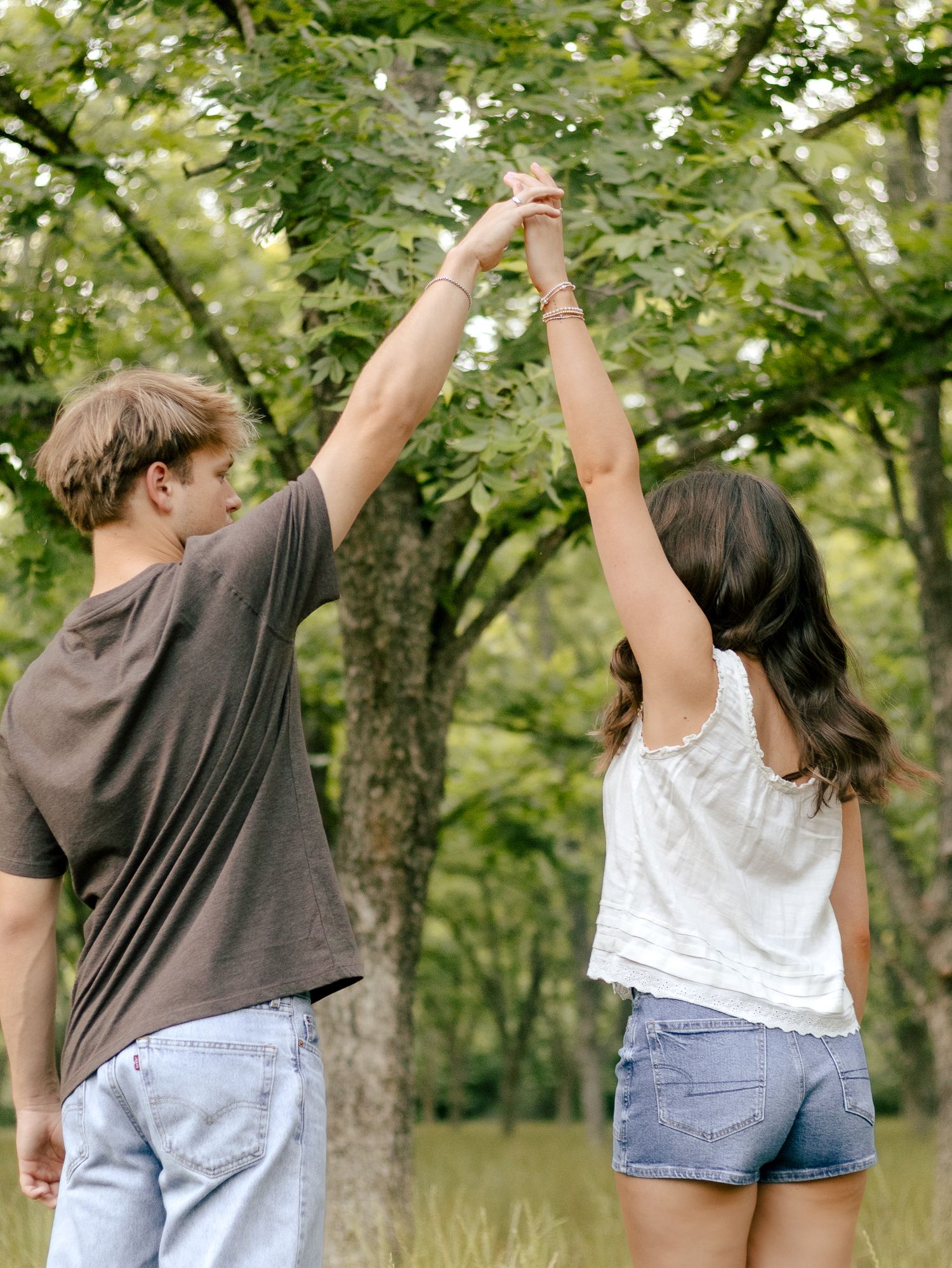 Young couple holding hands raised high while standing together under green trees in a sunny park, smiling in a romantic outdoor moment.