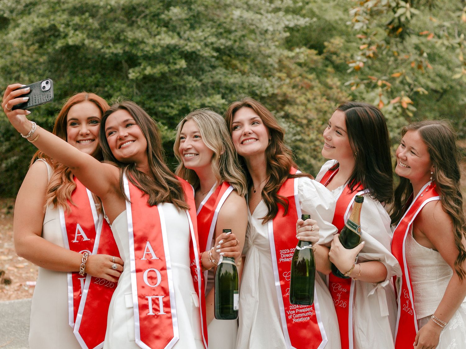 Group of smiling women in white dresses and red sorority sashes take a selfie while holding champagne bottles outdoors among trees.