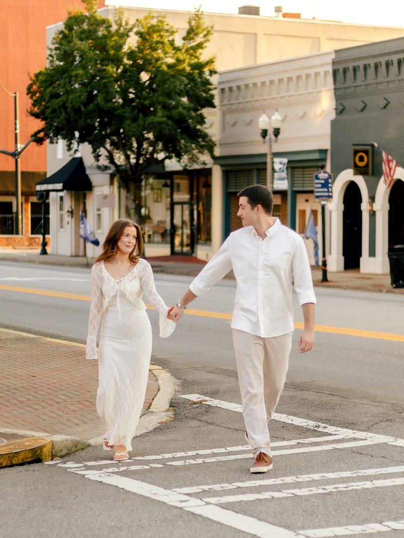 Engaged couple in matching white outfits holding hands and walking across a downtown crosswalk during an outdoor engagement photo session.