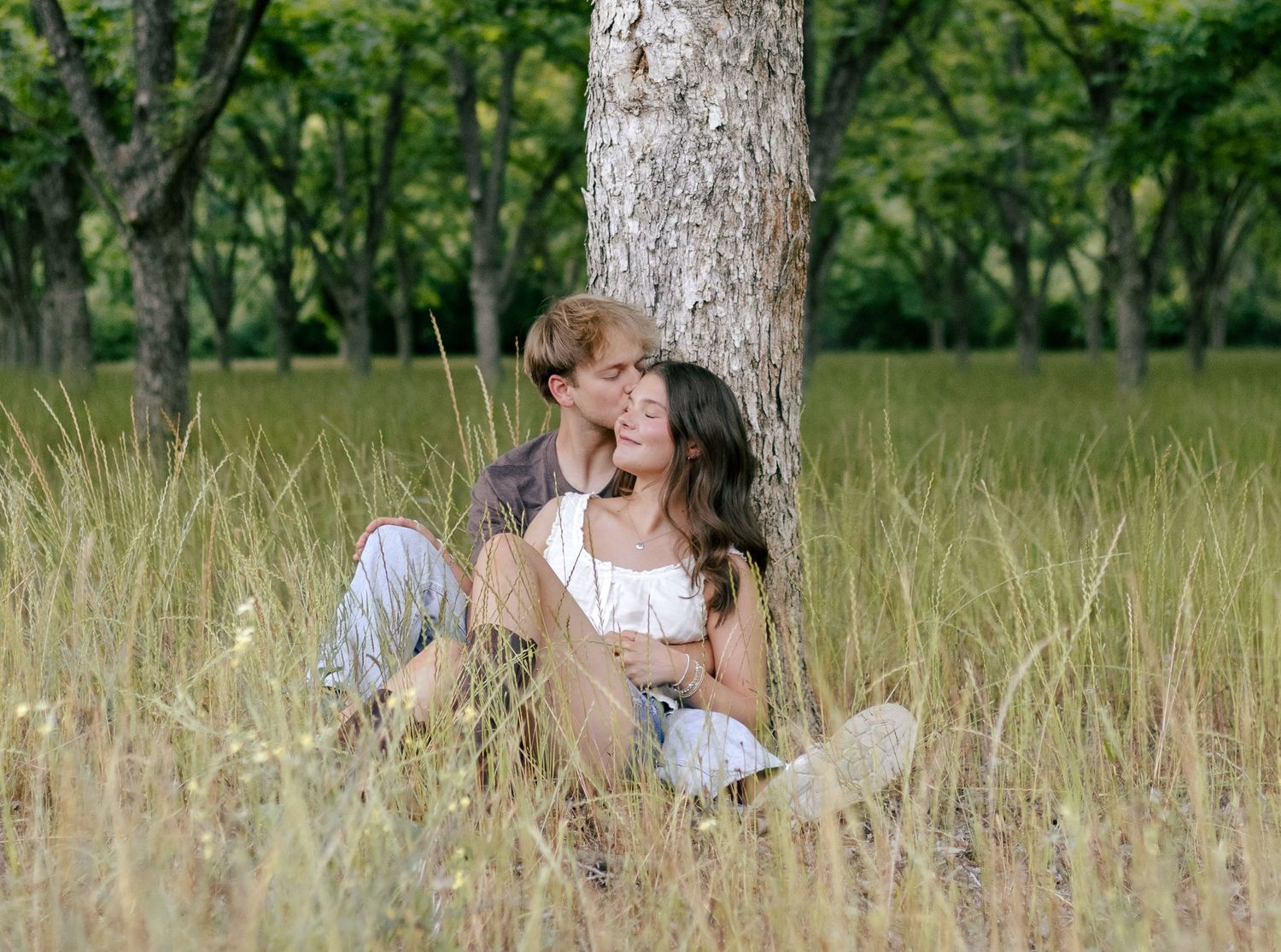 A couple sits close together in tall golden grass, leaning against a tree trunk in a sunlit orchard, sharing an intimate romantic moment.