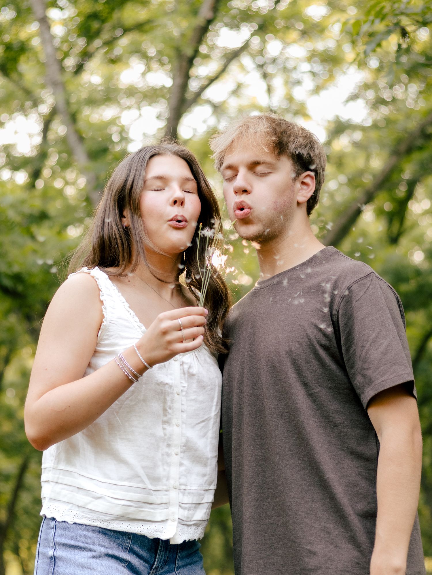 Young woman and man stand close together outdoors, blowing dandelion seeds into the air among green trees.