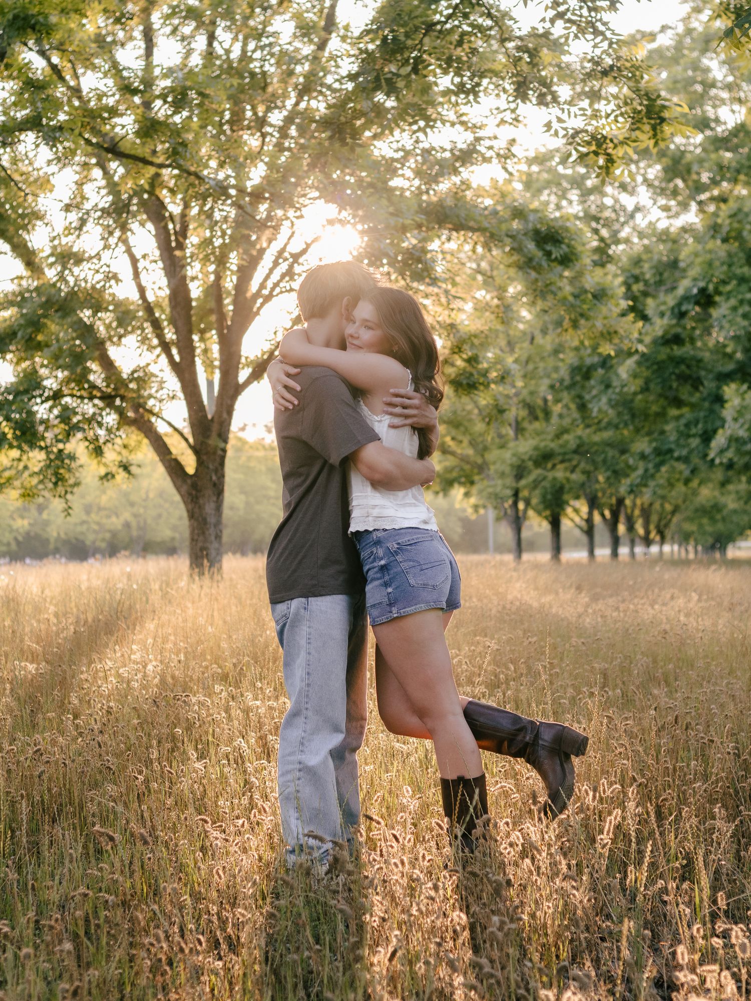 A couple embraces in a golden grass field at sunset, with the sun shining through tall trees in the background.