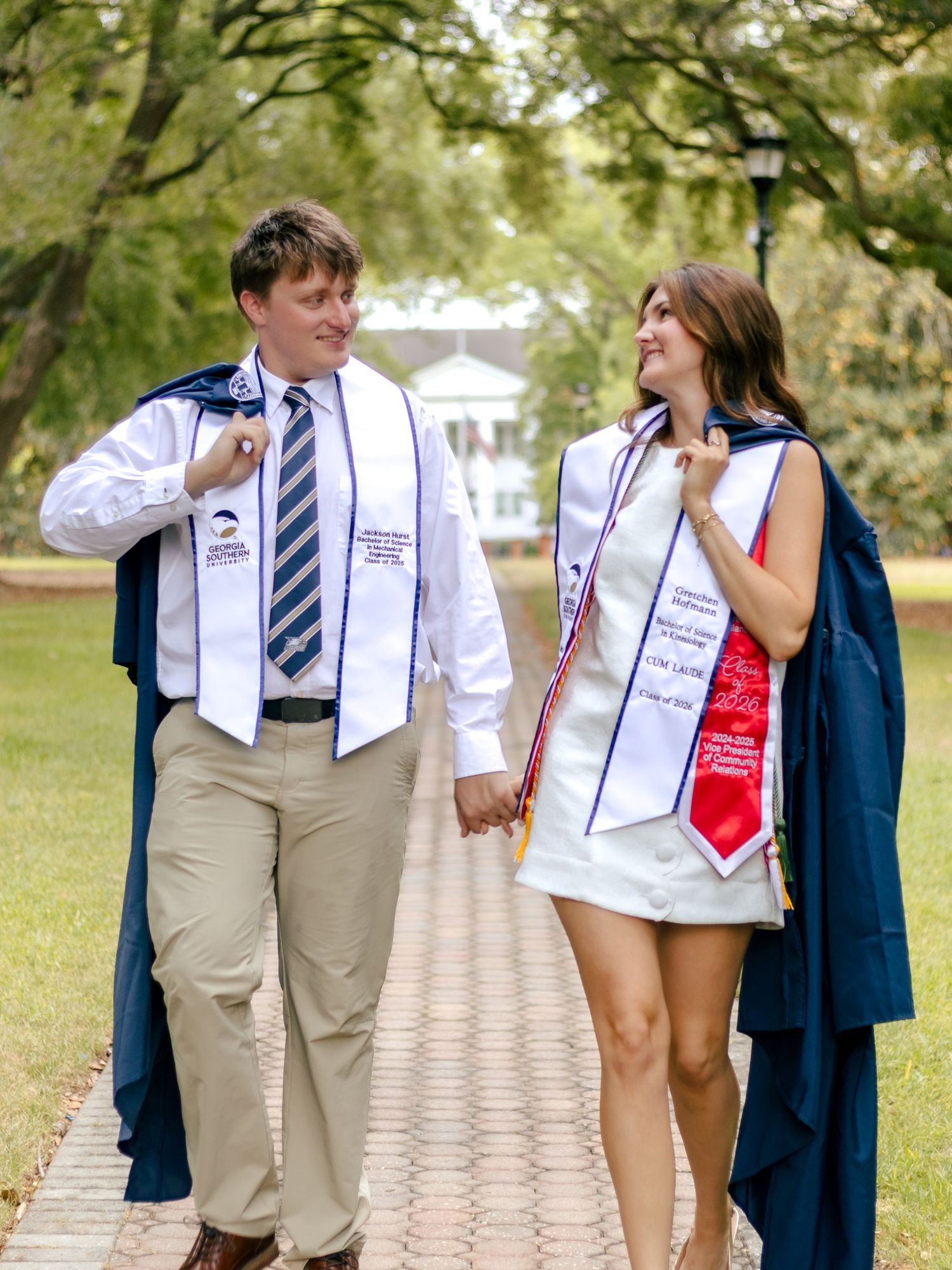 Two smiling graduates in white sashes and navy gowns hold hands while walking down a brick path on a tree-lined campus.