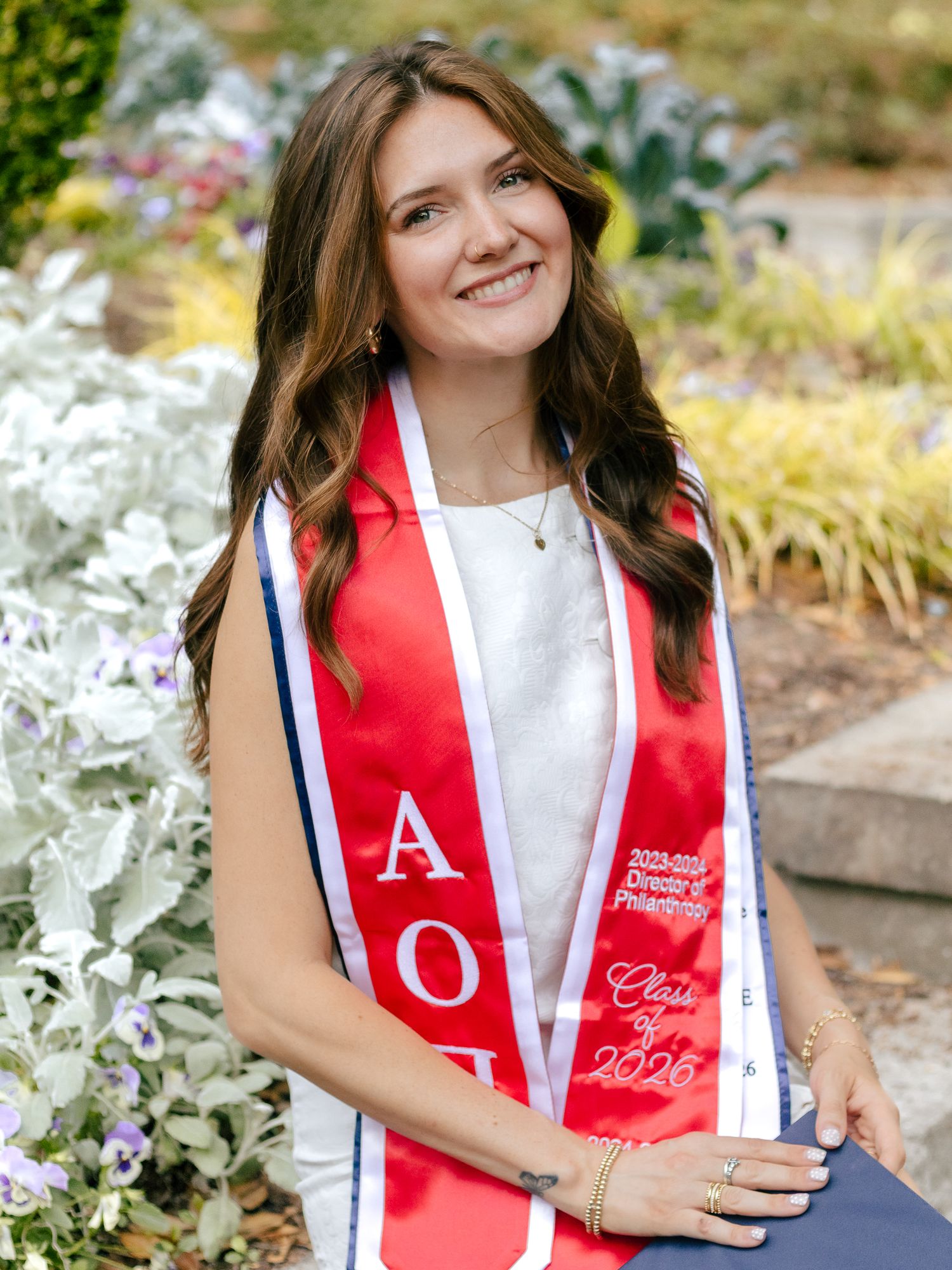 Smiling young woman with long brown hair wears a red and white Alpha Omicron Pi graduation sash while posing outdoors in a garden.