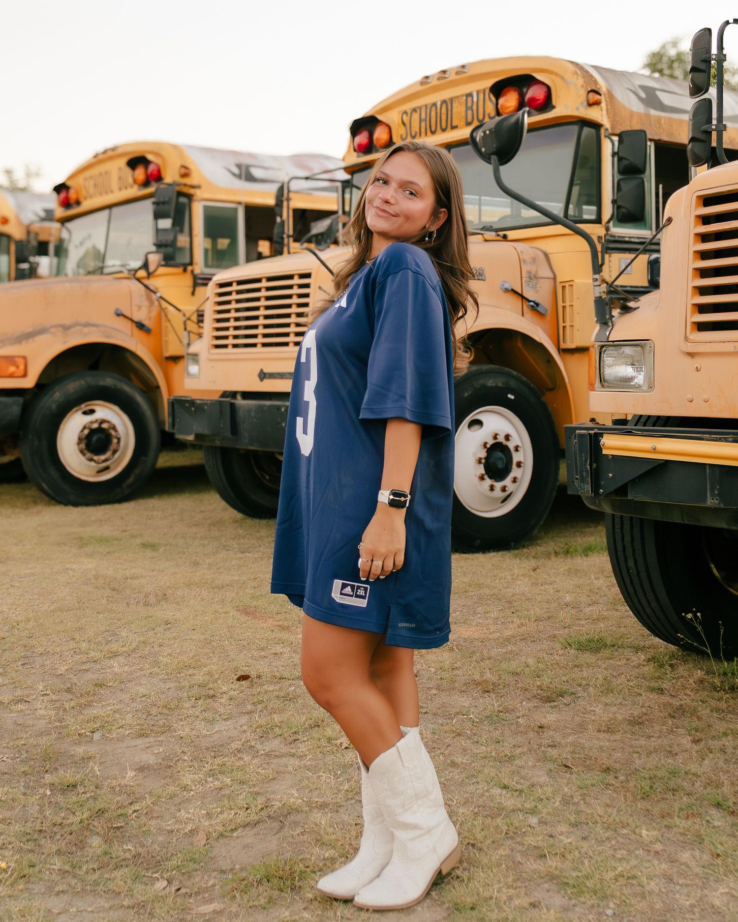 Young woman in a blue number 3 jersey dress and white cowboy boots posing in front of a row of yellow school buses.