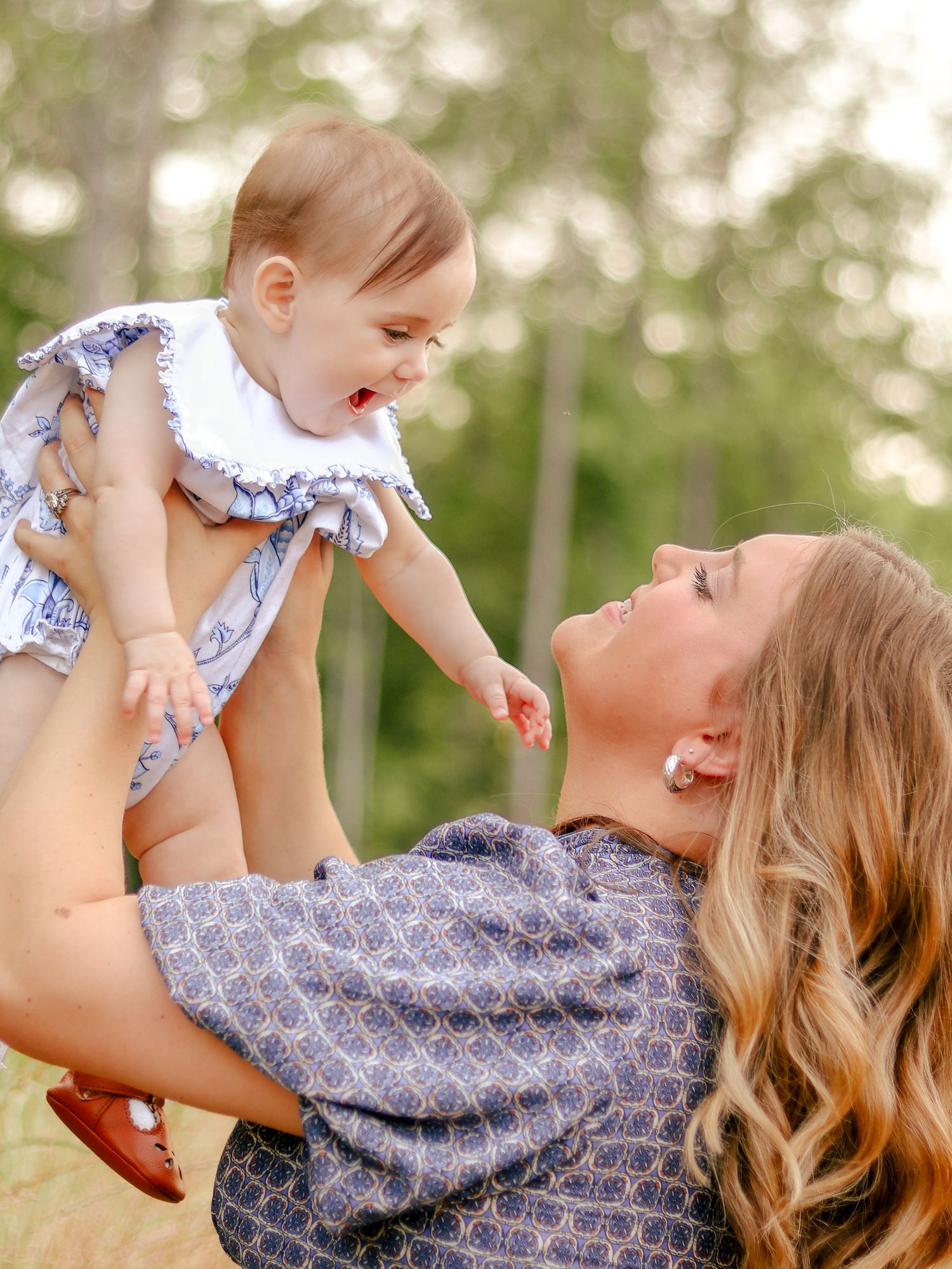 Smiling woman with long wavy hair lifts a laughing baby in a white ruffled outfit into the air outdoors among soft green trees.