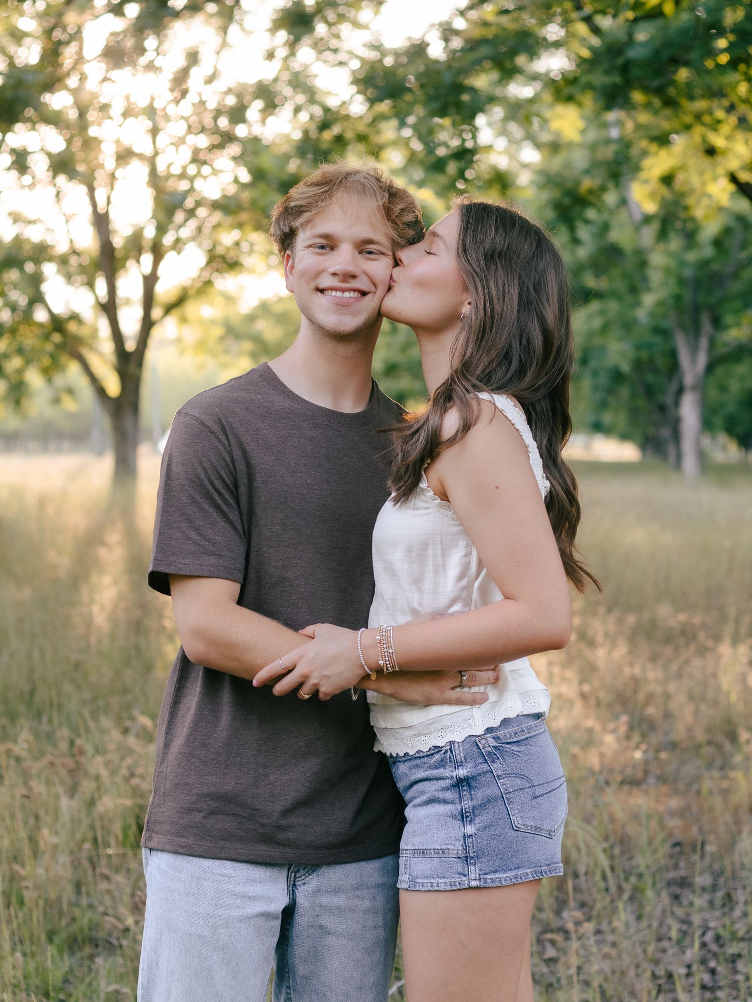 A smiling young man in a brown t-shirt is kissed on the cheek by a young woman in a white top and denim shorts in a sunlit grassy field.