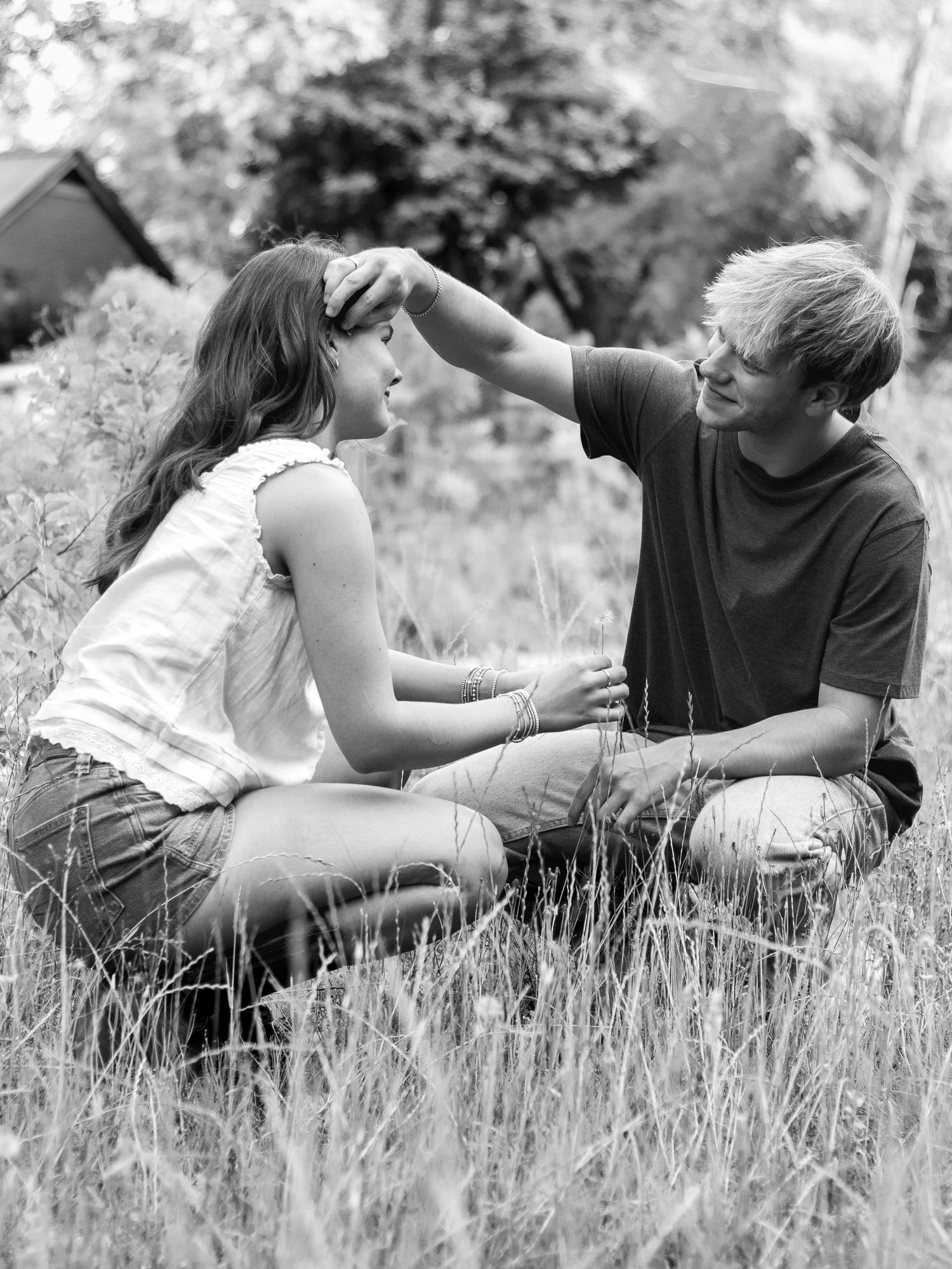 Black and white photo of a young couple kneeling face to face in tall grass as he gently touches her hair.