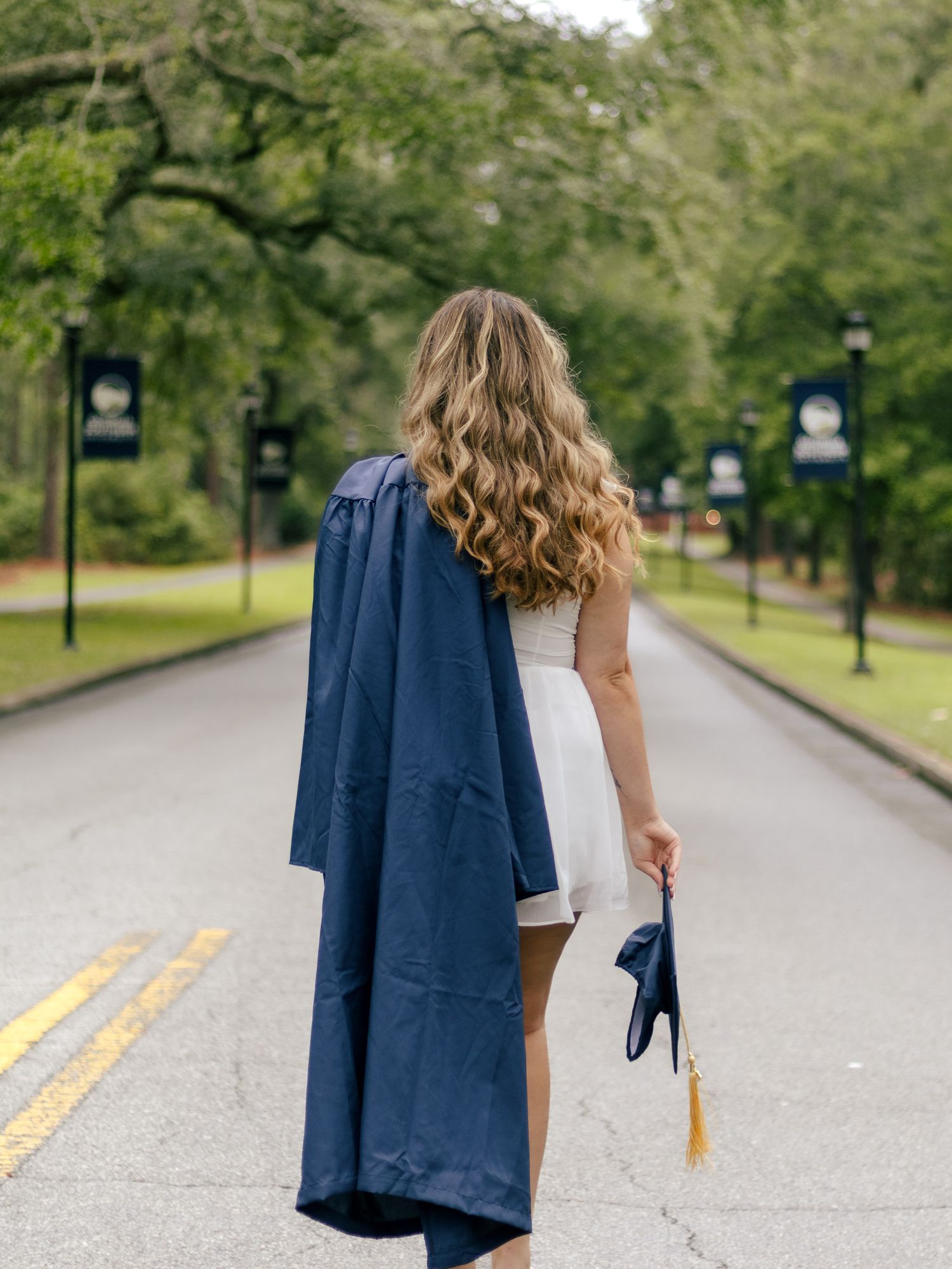 Graduate with long wavy hair walks down a tree-lined campus road holding a navy gown and cap with a gold tassel.