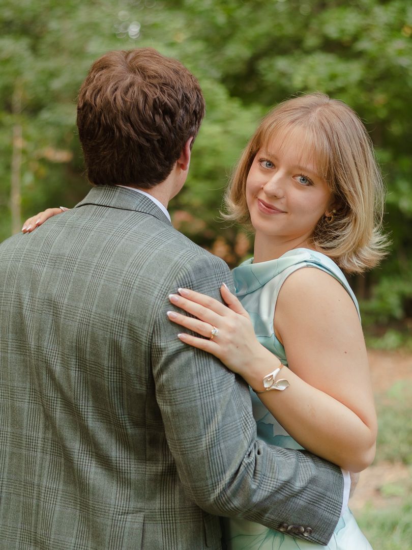 A smiling woman in a light blue dress rests her ring-adorned hand on a man's shoulder as they embrace in a wooded setting.