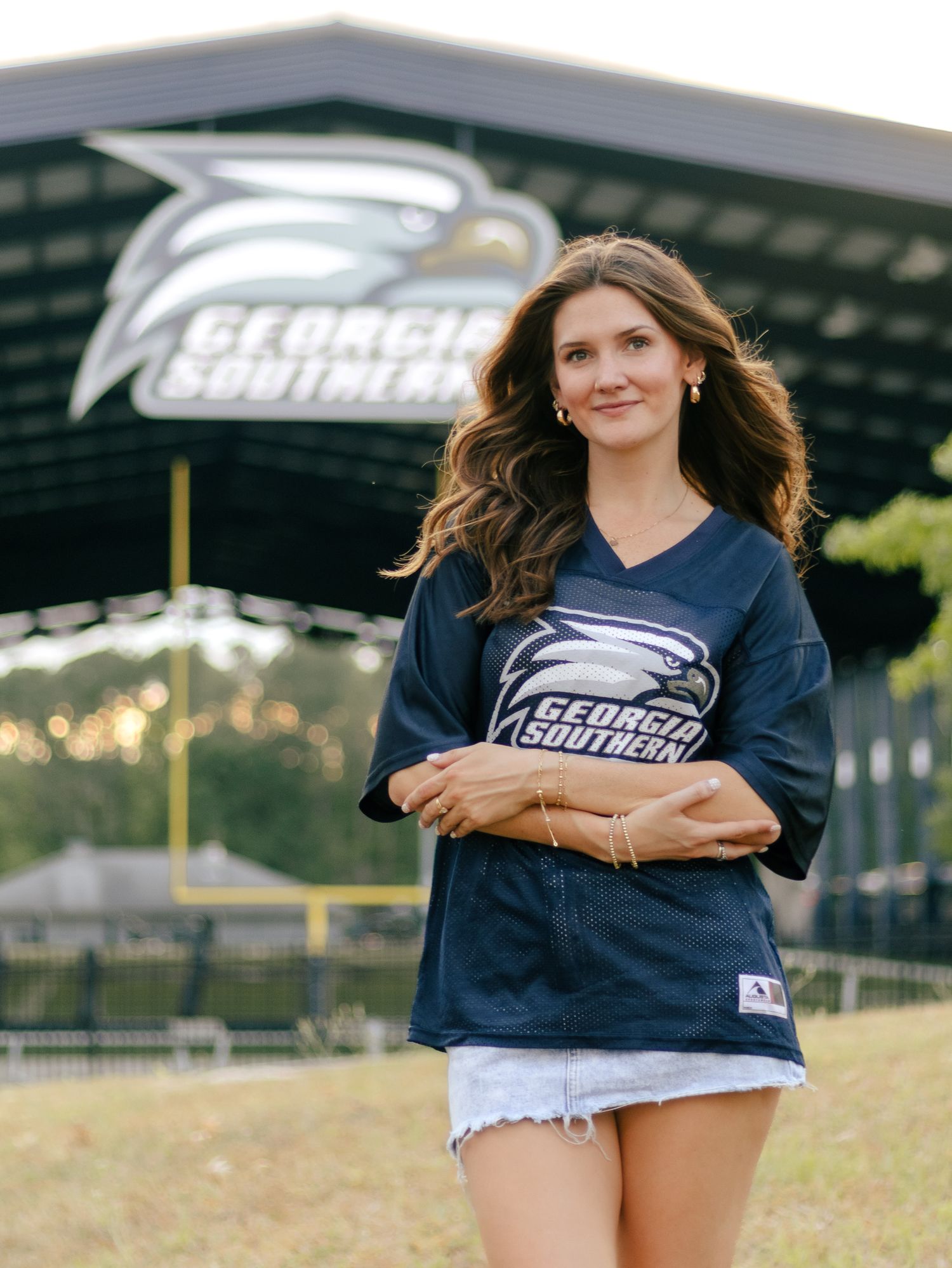 Woman with long wavy hair in a navy Georgia Southern jersey and denim shorts stands arms crossed outside the stadium.