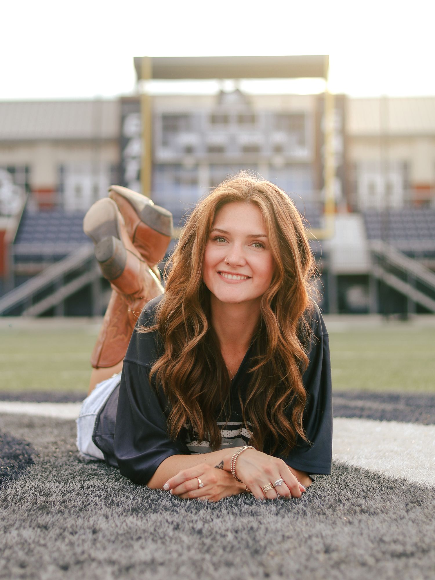 Smiling young woman with long wavy auburn hair and cowboy boots lies on artificial turf in a football stadium at sunset.