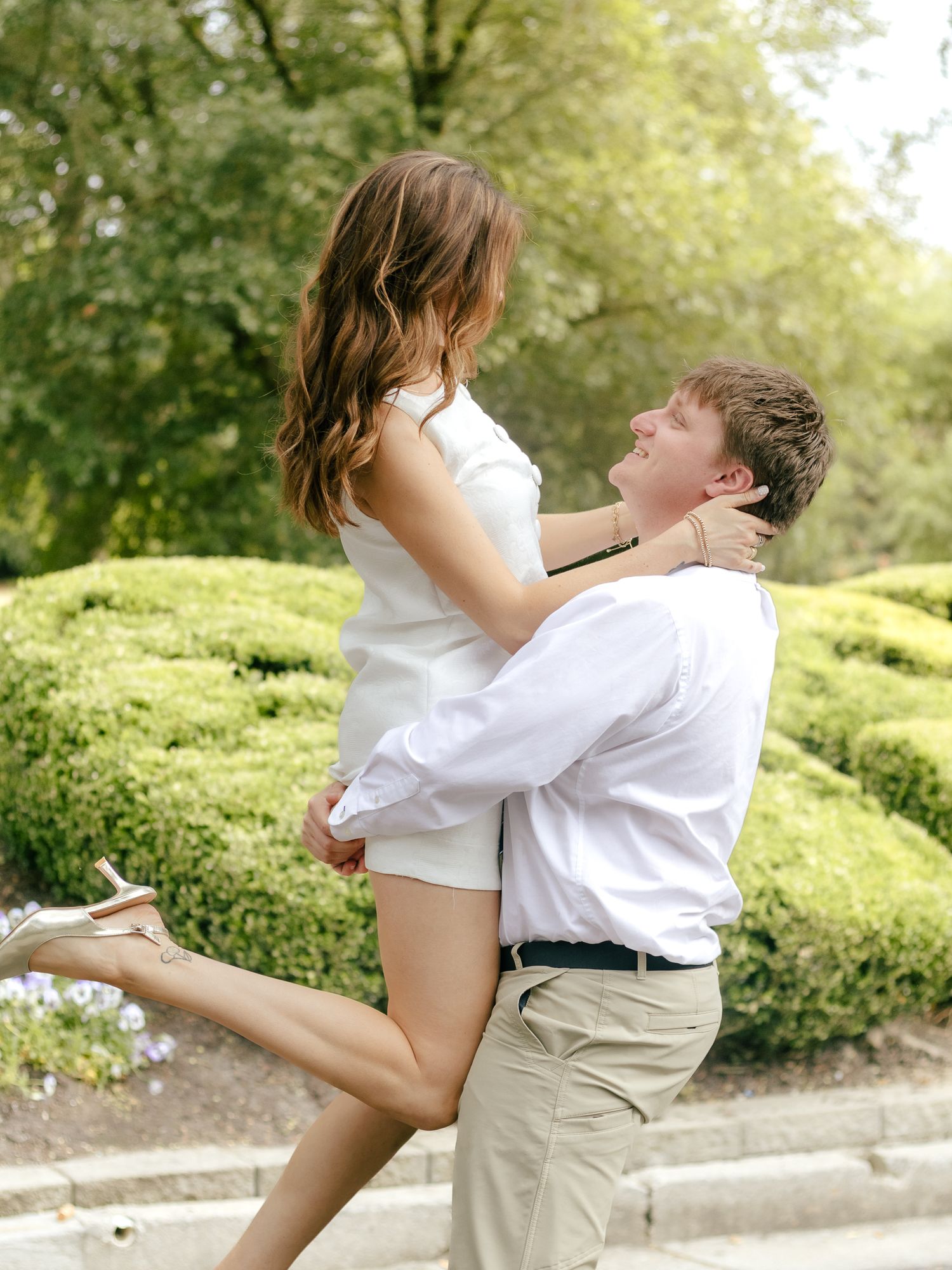 Couple embracing outdoors as a man in a white shirt lifts a smiling woman in a white dress in front of trimmed green hedges.
