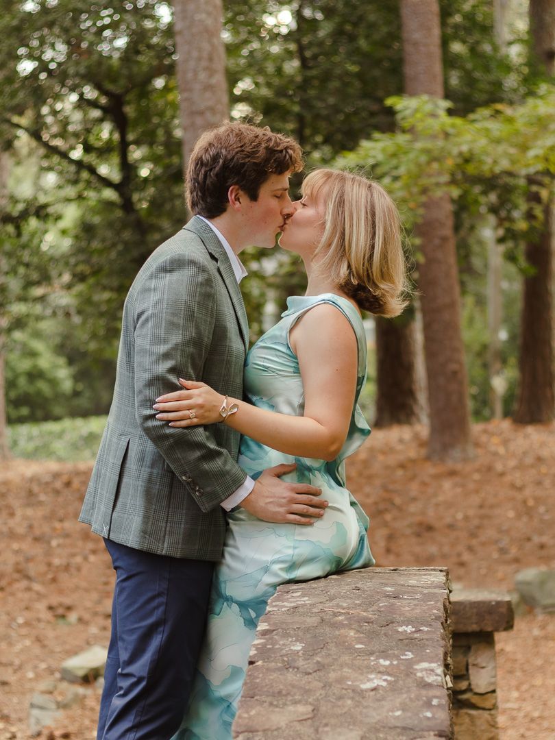 Couple sharing a kiss in a wooded park, he in a gray blazer and she in a teal dress, seated on a stone ledge among tall pine trees.