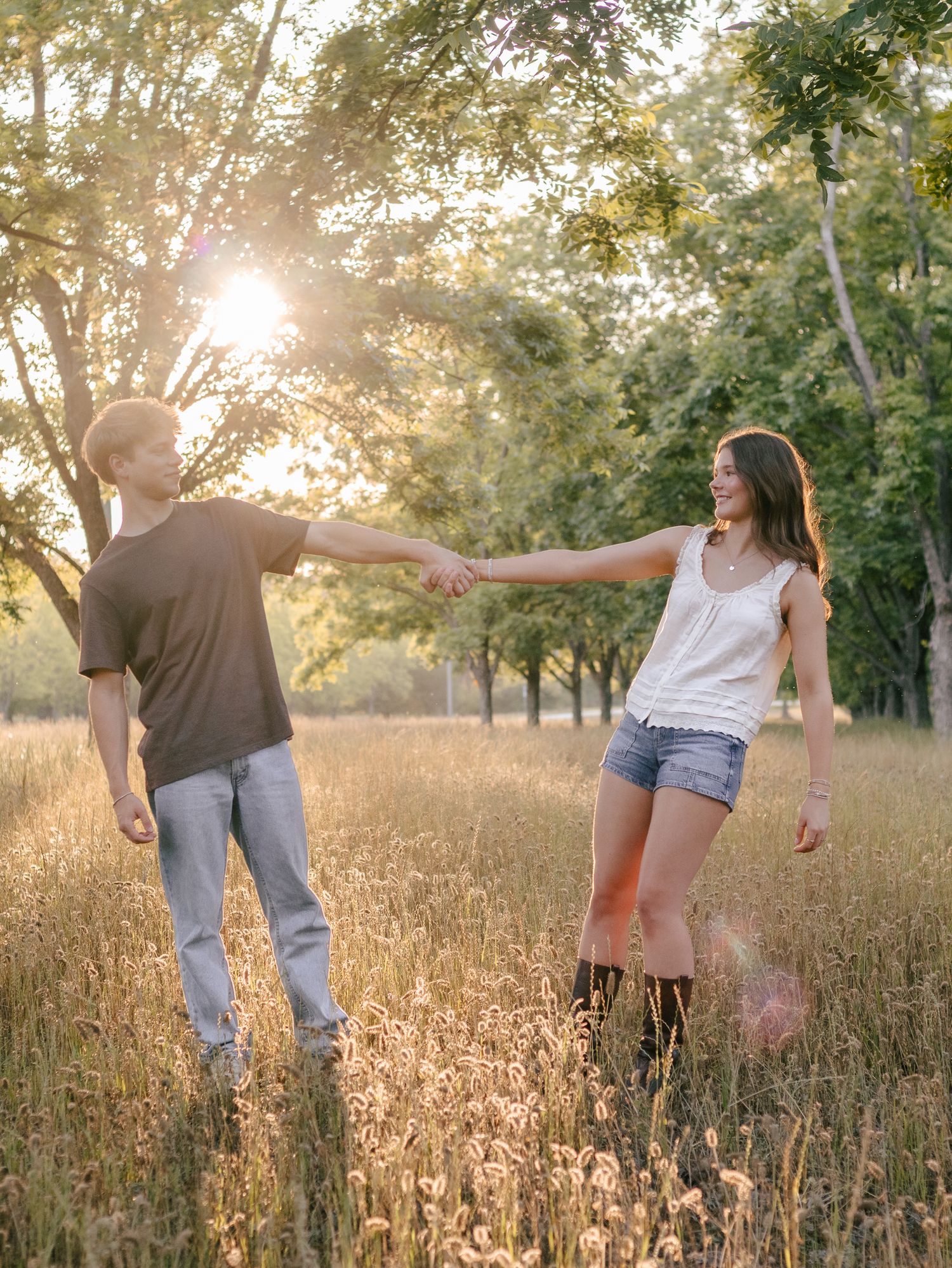 Young couple holding hands and smiling while walking through a golden grassy field with trees and warm sunlight glowing behind them.