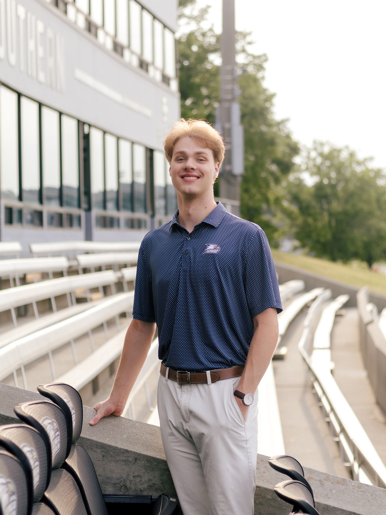 Smiling young man in a navy polo and khaki pants stands beside empty stadium seats with a press box and trees in the background.