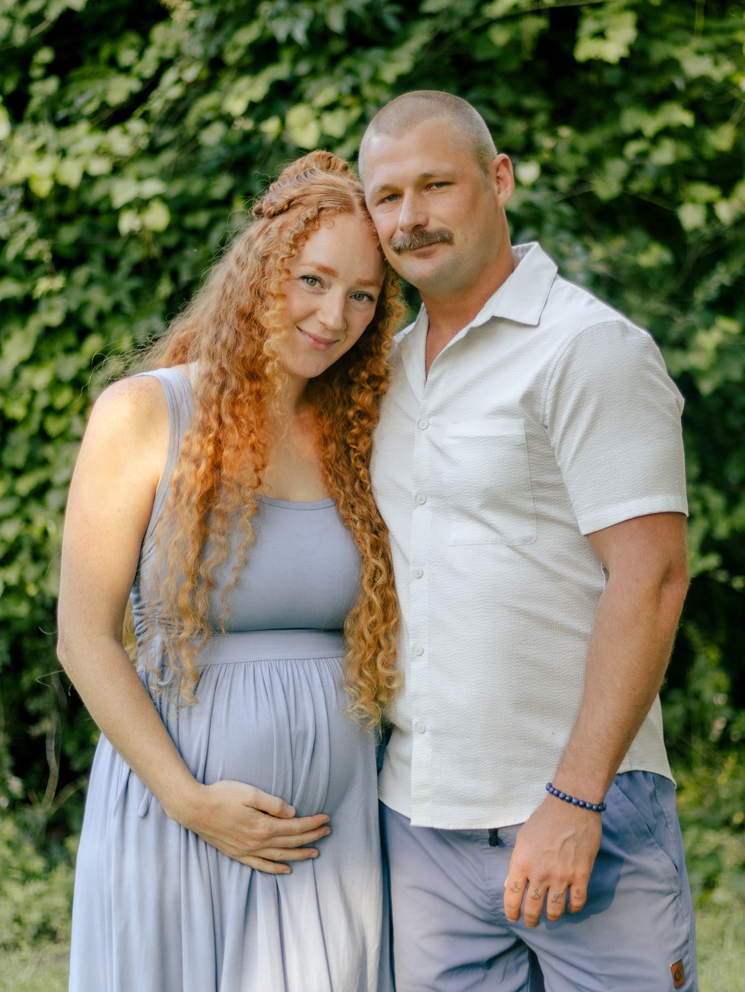 Expecting couple embracing outdoors, a long-haired woman in a blue maternity dress cradling her bump beside a smiling man in a white shirt.