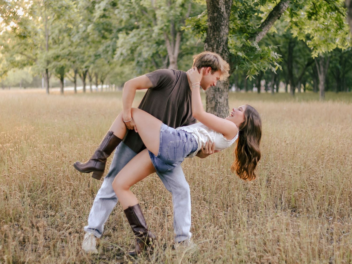 Couple dancing in a grassy field as a man dips a smiling woman in a playful tango pose, surrounded by tall trees in warm sunlight.