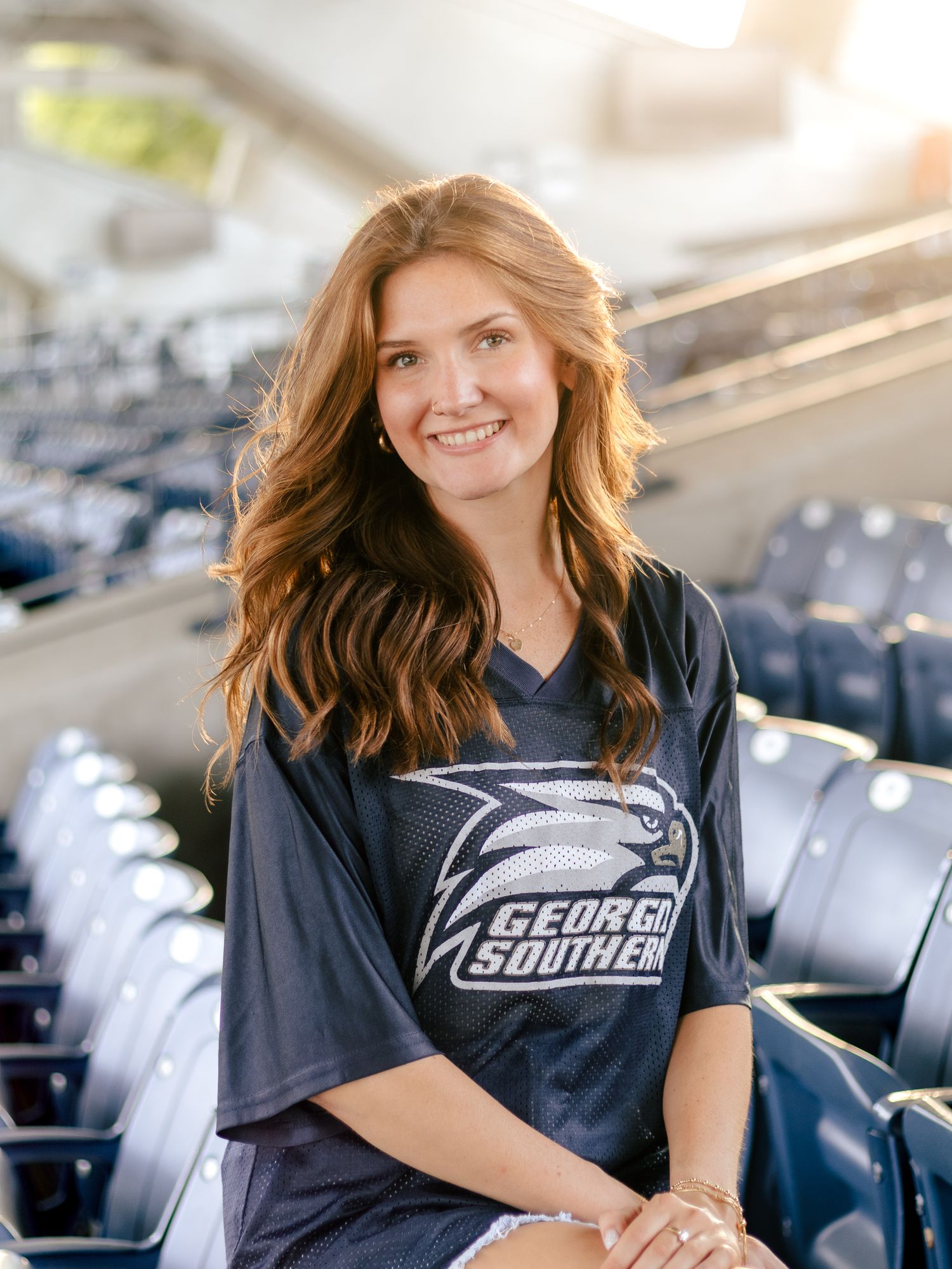 Smiling woman with long auburn wavy hair wears a navy Georgia Southern jersey while sitting in empty stadium seats.