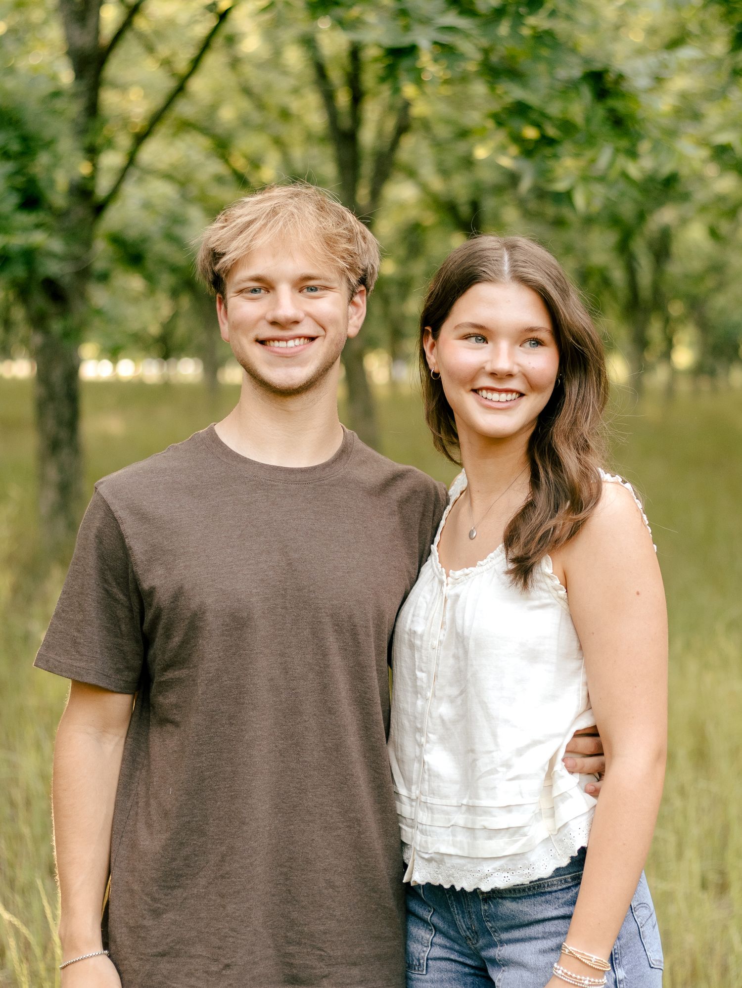 A smiling young man in a brown t-shirt and a young woman in a white lace top pose together outdoors in a sunlit grassy field with trees.
