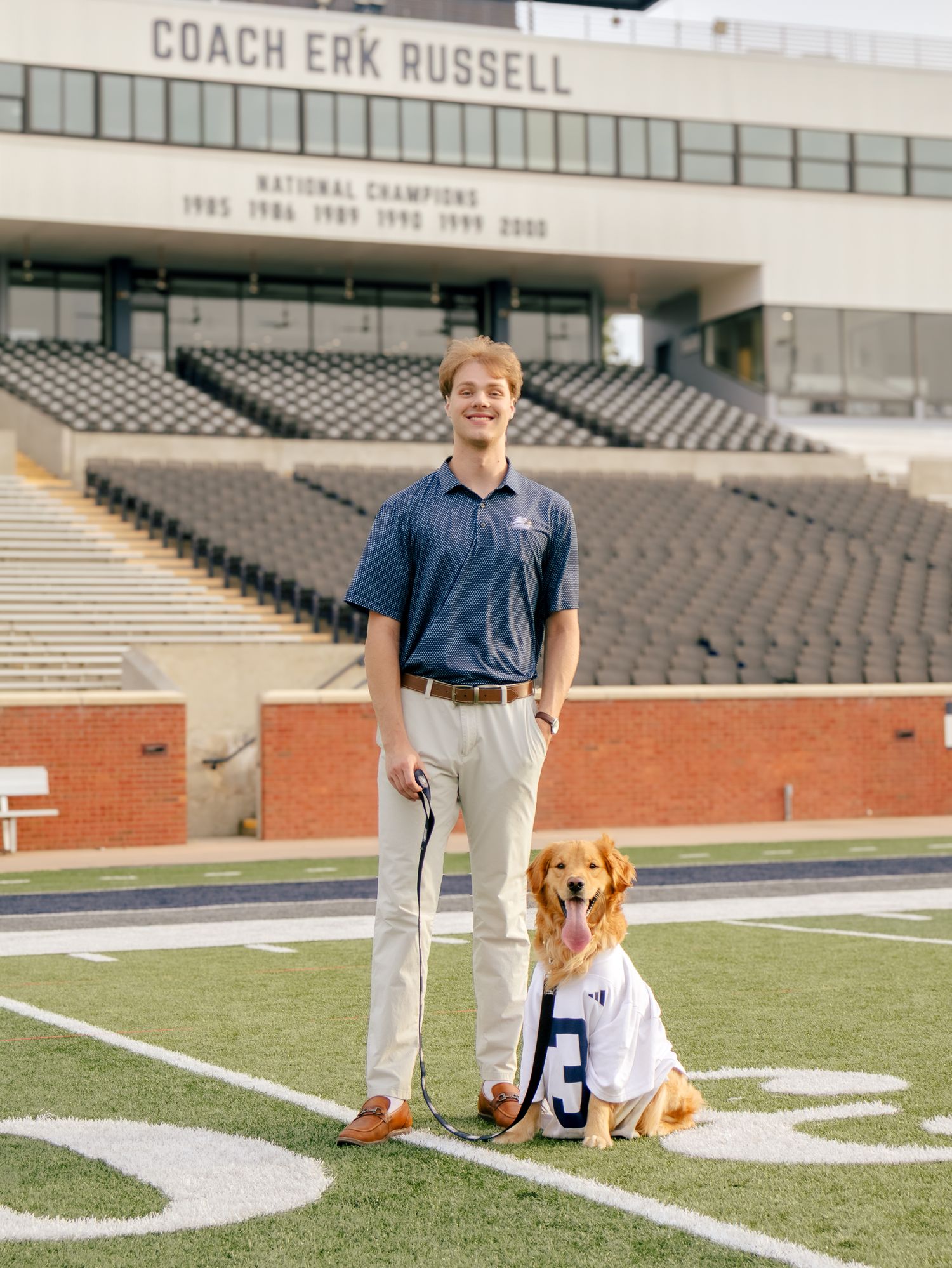Young man in a navy polo and khakis stands on a football field holding the leash of a golden retriever wearing a jersey.