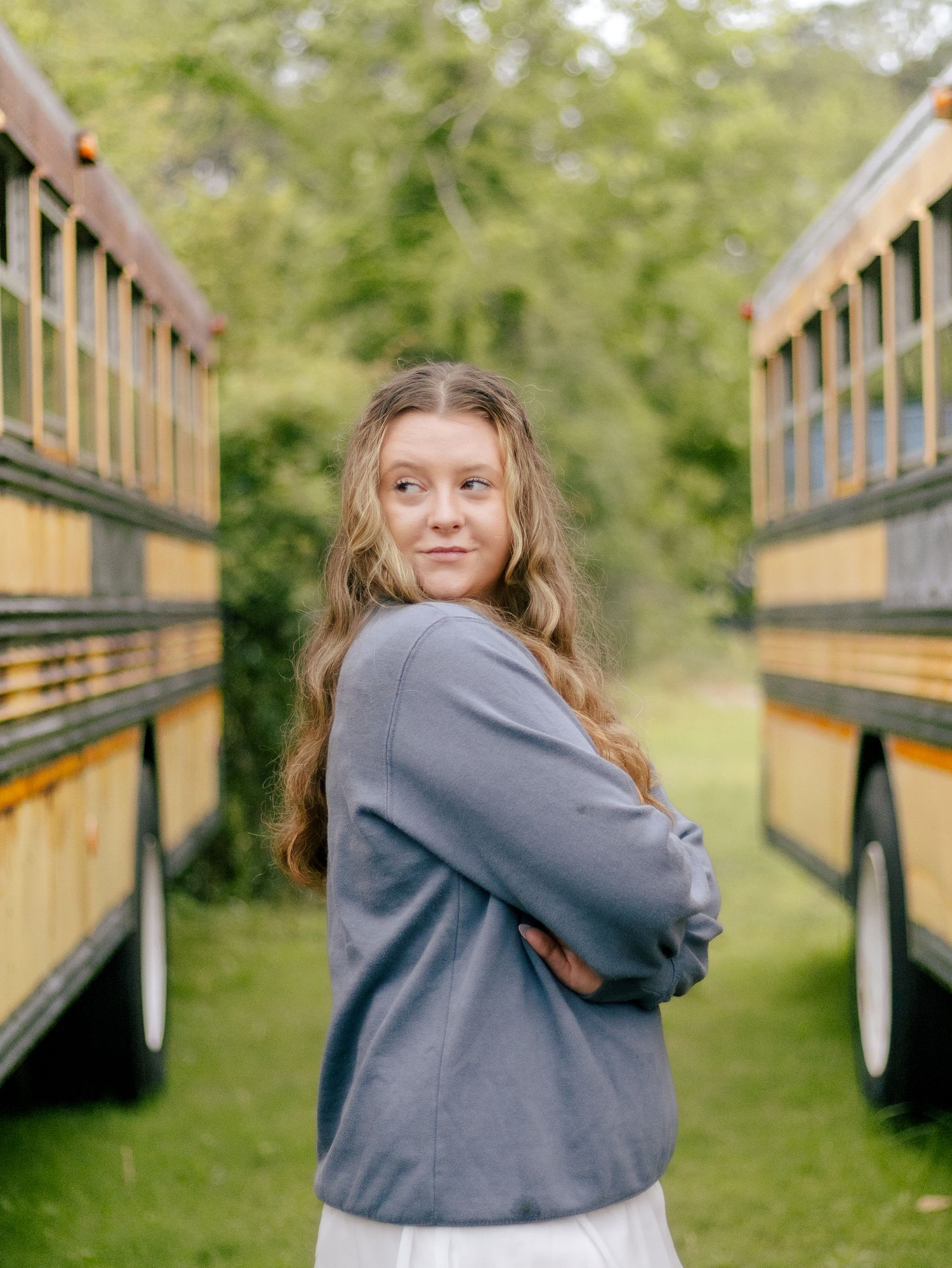 Young woman with long wavy hair in a gray sweatshirt looks over her shoulder between two yellow school buses on a grassy path.