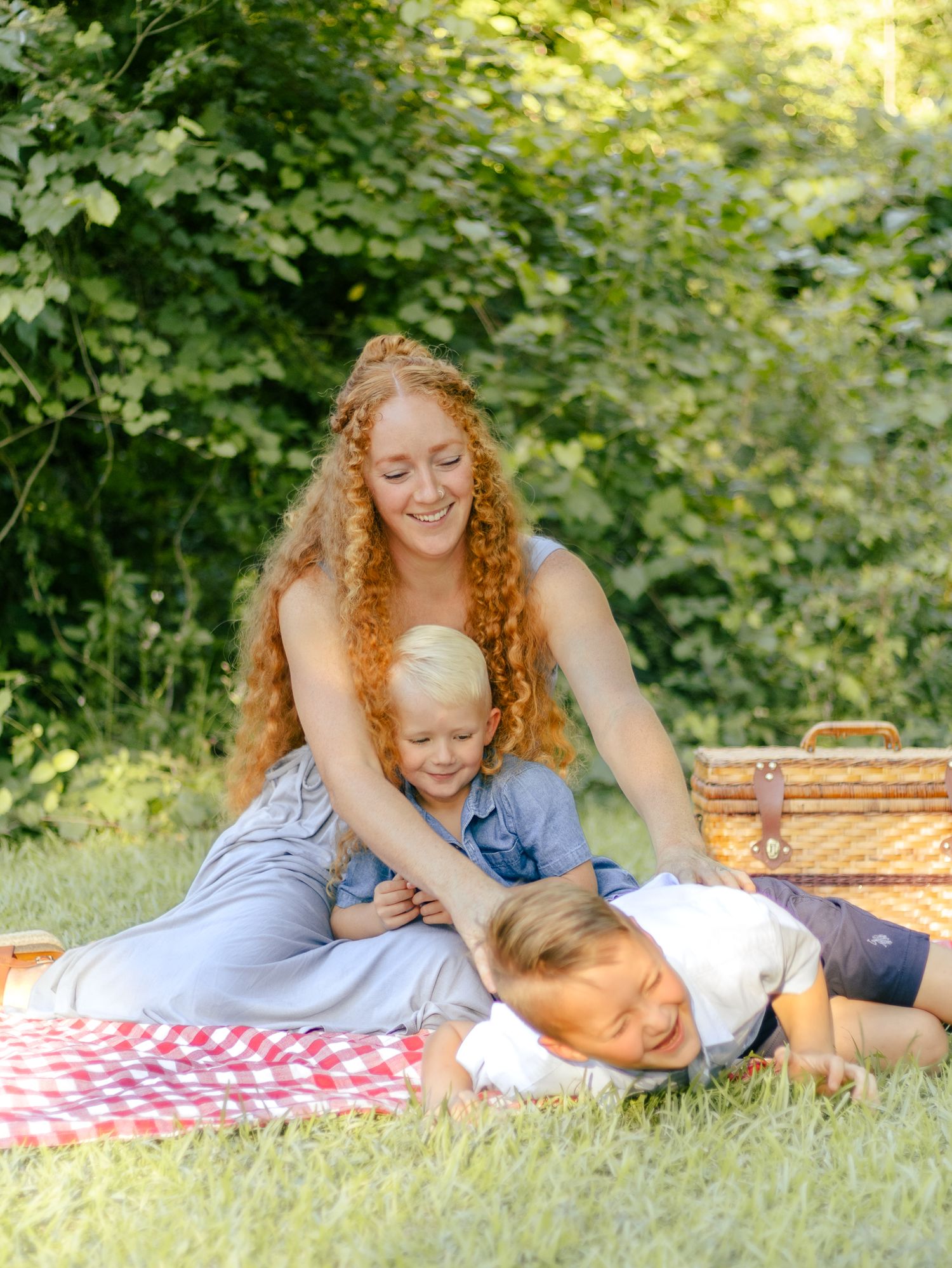 Smiling woman with long curly red hair sits on a red gingham picnic blanket hugging two laughing young boys beside a wicker basket.