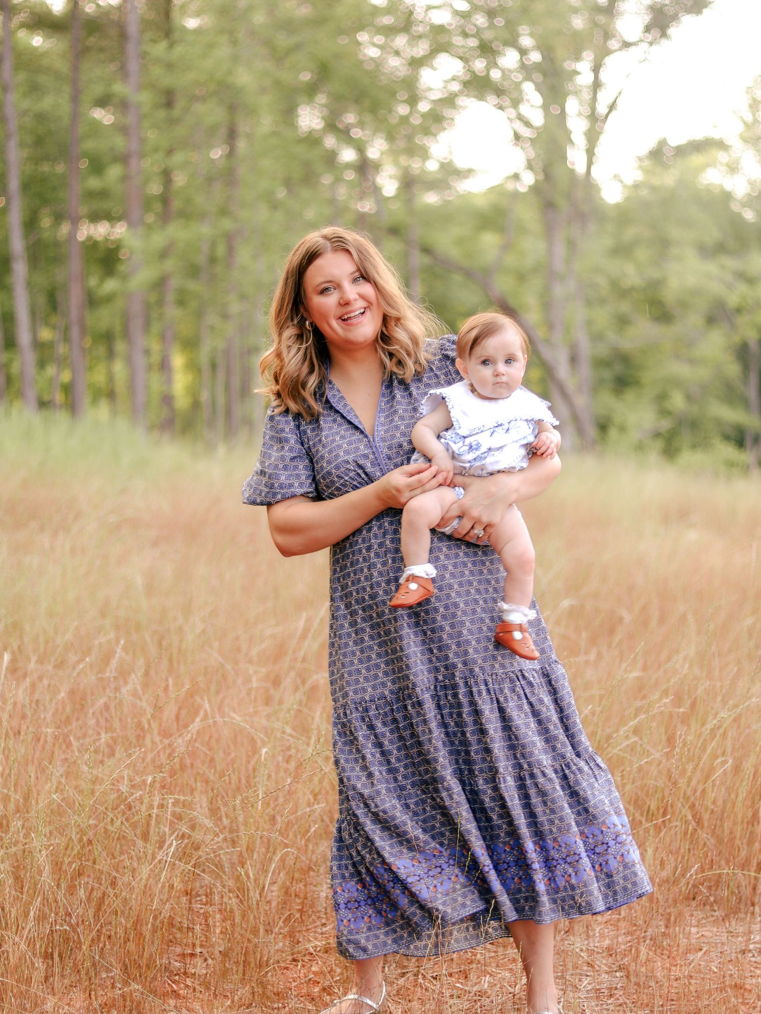 Smiling woman in a blue patterned dress holding a baby in a white top and orange shoes in a golden grass field with tall trees.