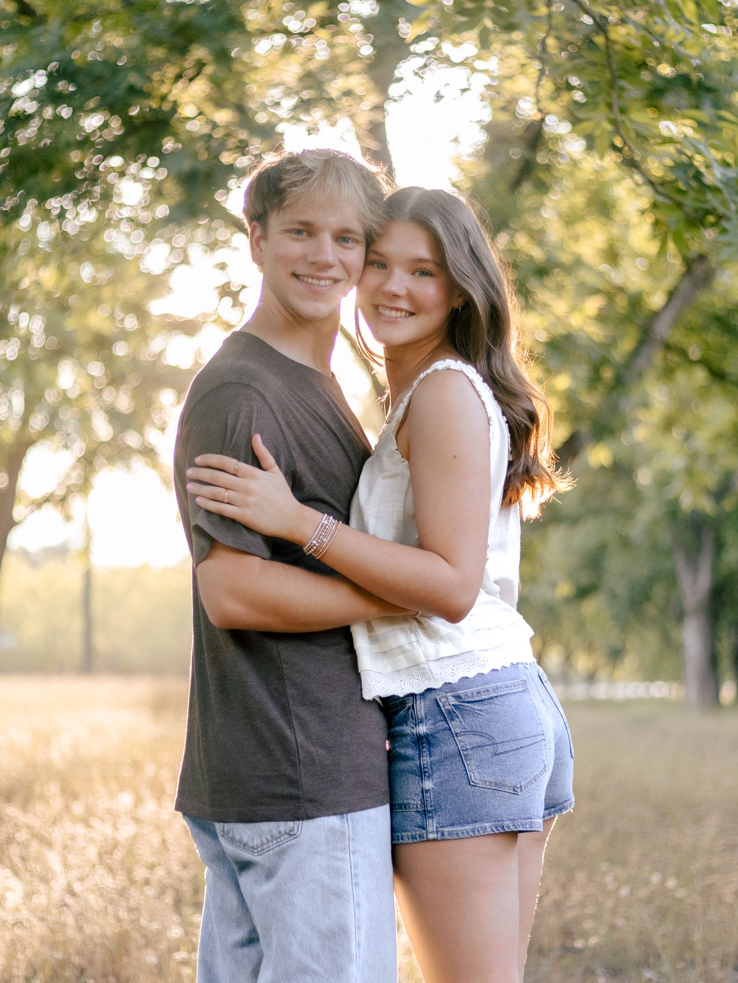 A smiling young couple embraces outdoors in golden evening light, standing together in a grassy field with trees behind them.