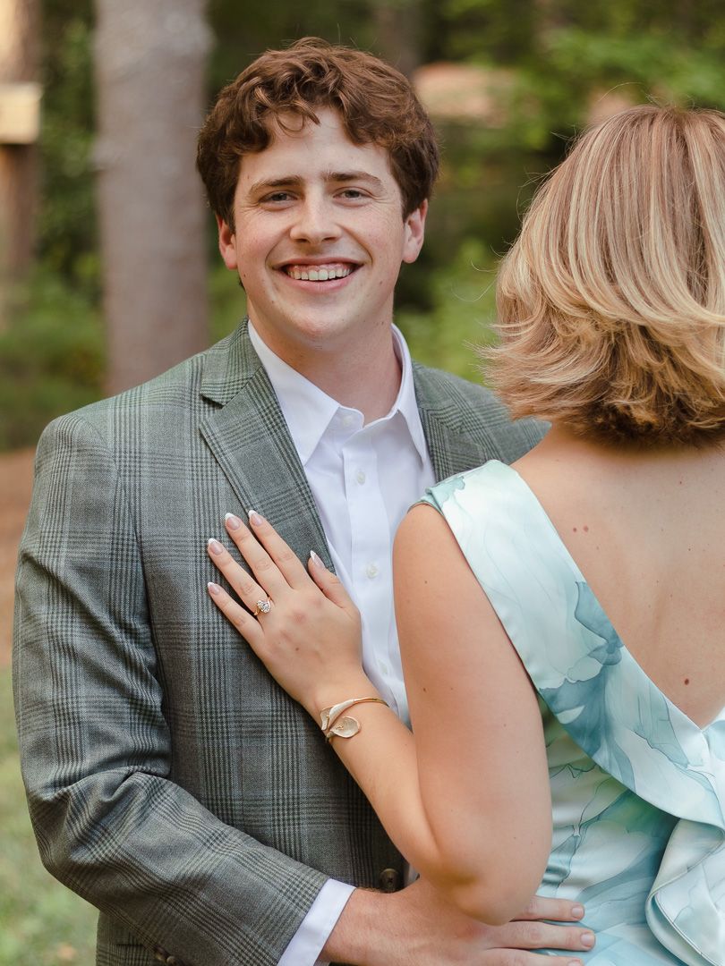 A smiling young man in a gray plaid blazer and white shirt stands in a forest as a woman's hand with a ring rests on his chest.