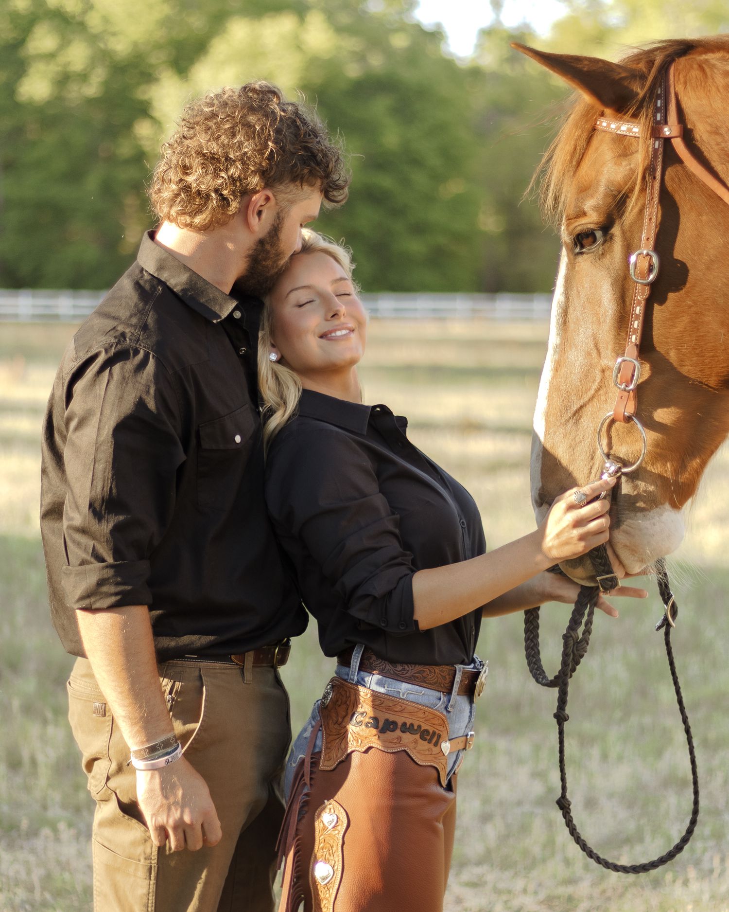 A smiling couple in western wear embraces beside a chestnut horse in a sunny field, the woman gently petting its nose.