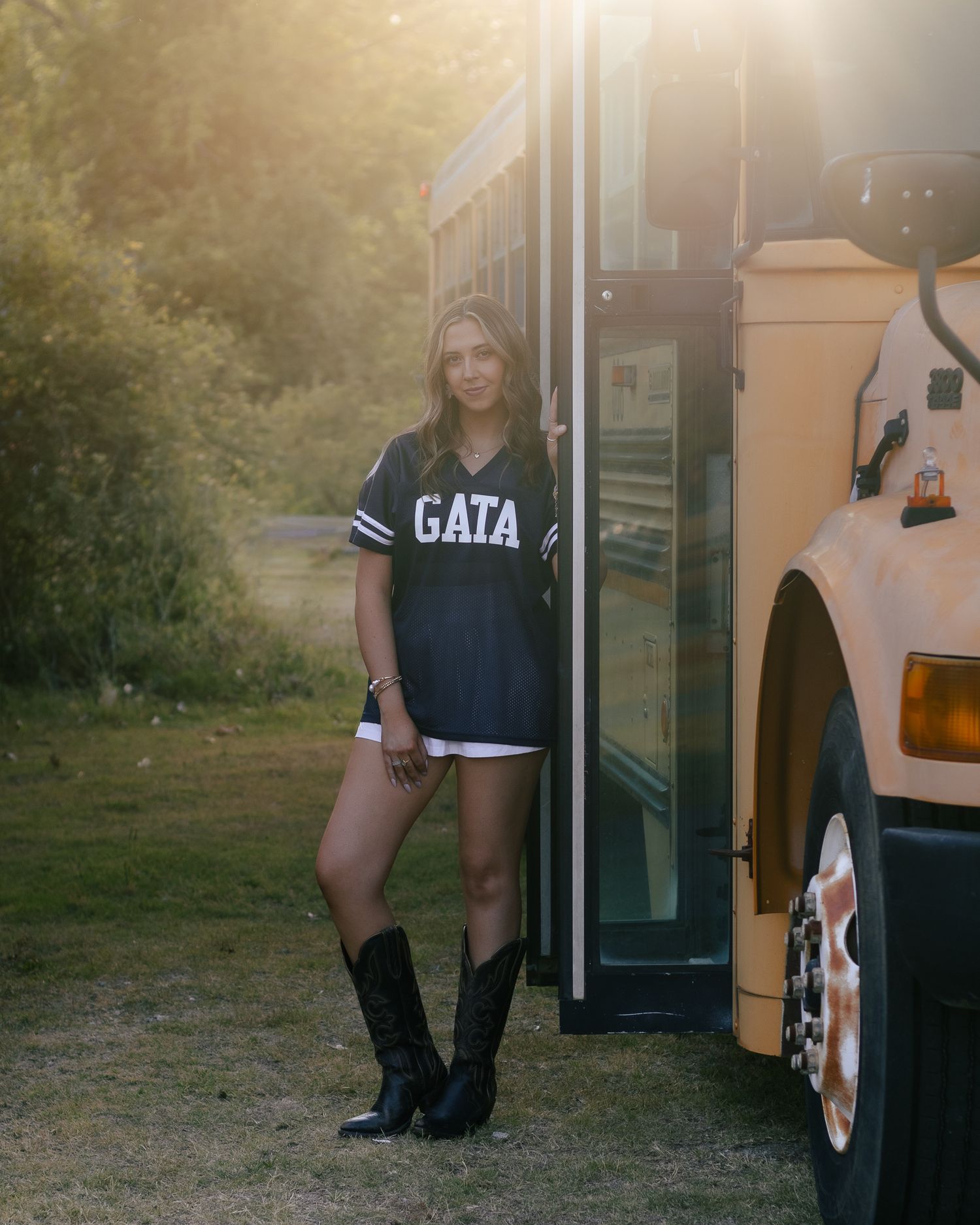 Young woman in a navy GATA jersey, white shorts and black cowboy boots leans against a yellow school bus door in warm golden sunlight.