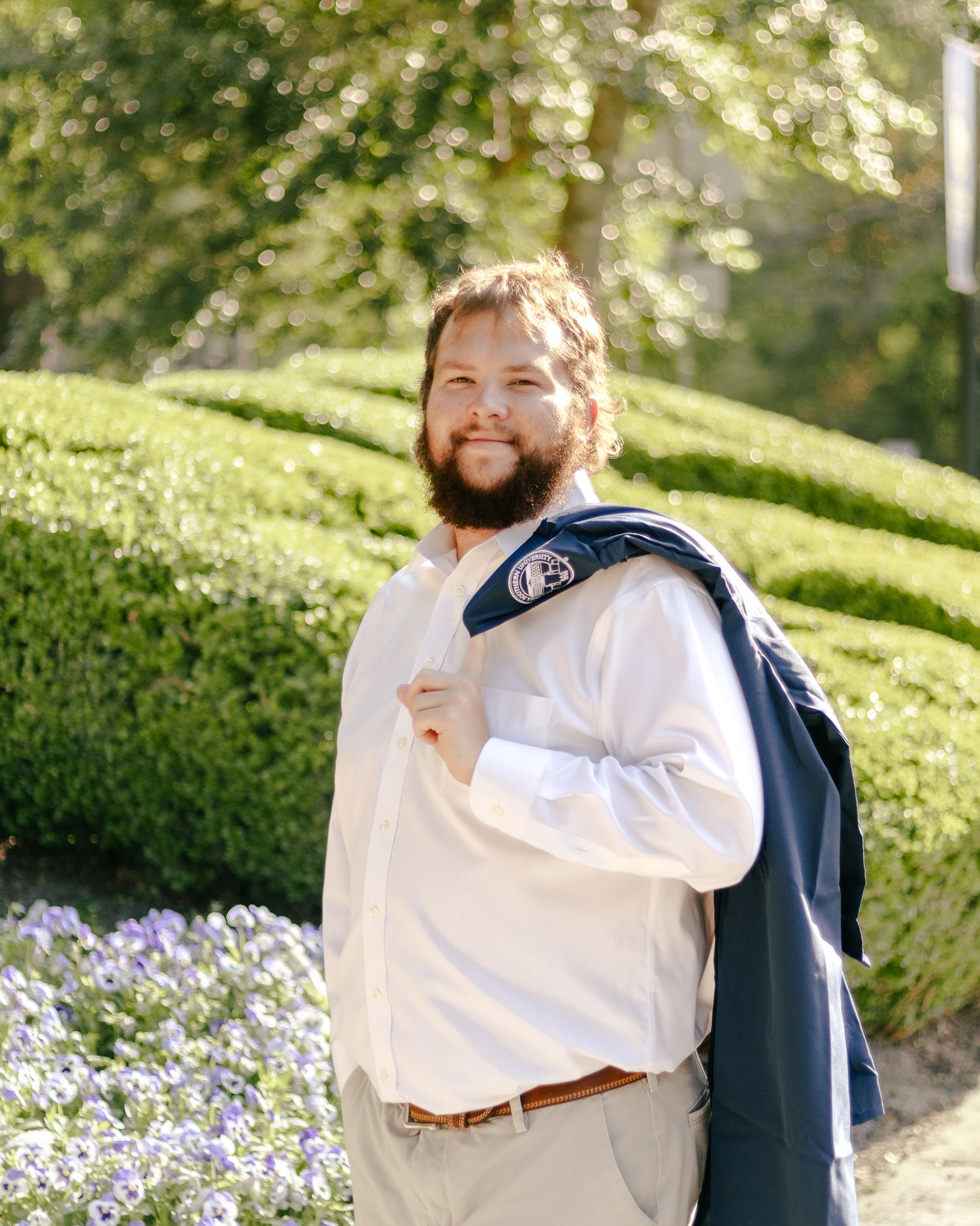 Smiling bearded man in a white dress shirt holds a navy jacket over his shoulder while standing near trimmed hedges and purple flowers.