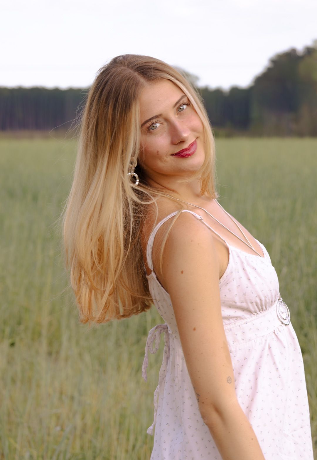 Smiling woman with long blonde hair wearing a white sundress poses in a green grassy field with trees in the background.