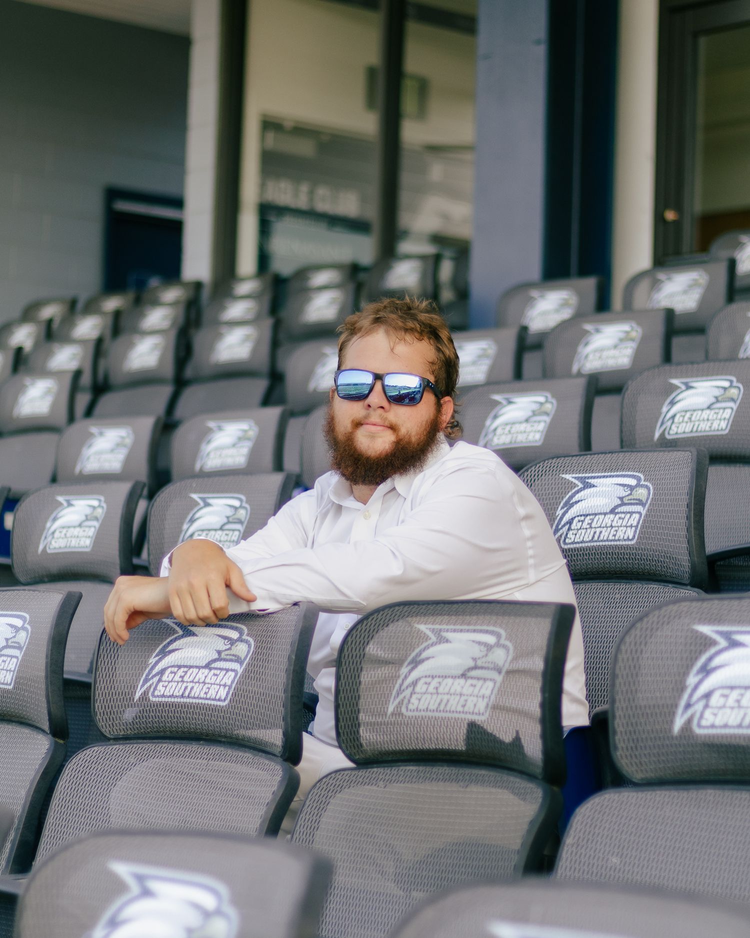 Bearded man in sunglasses and a white shirt sits among gray Georgia Southern stadium seats with eagle logos.
