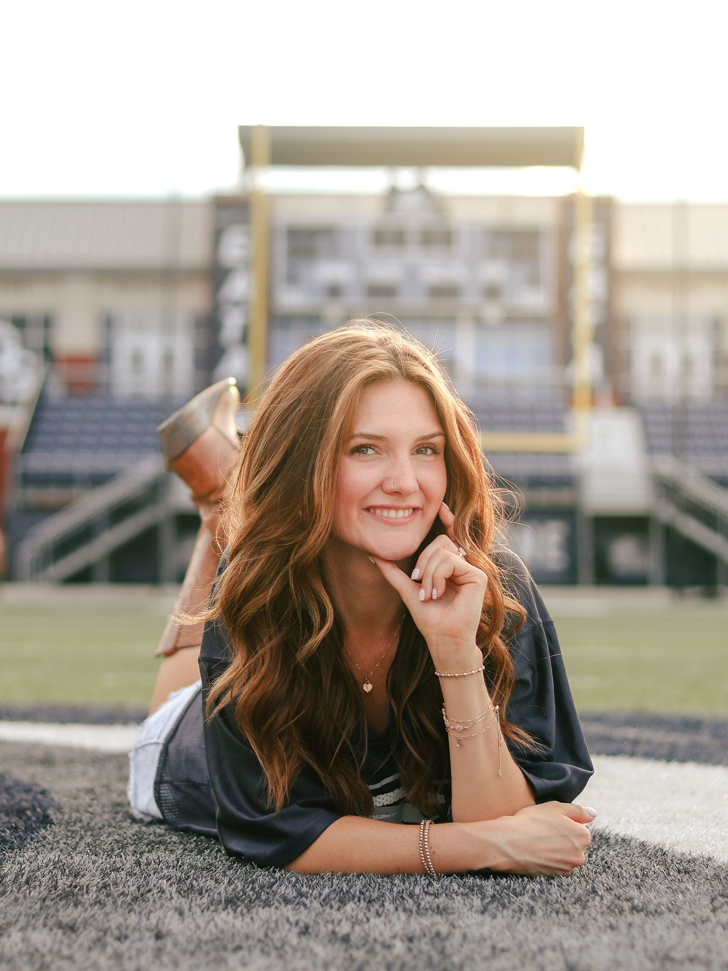 Smiling young woman with long wavy auburn hair lies on a turf field with her hand at her chin, stadium bleachers softly blurred behind her.