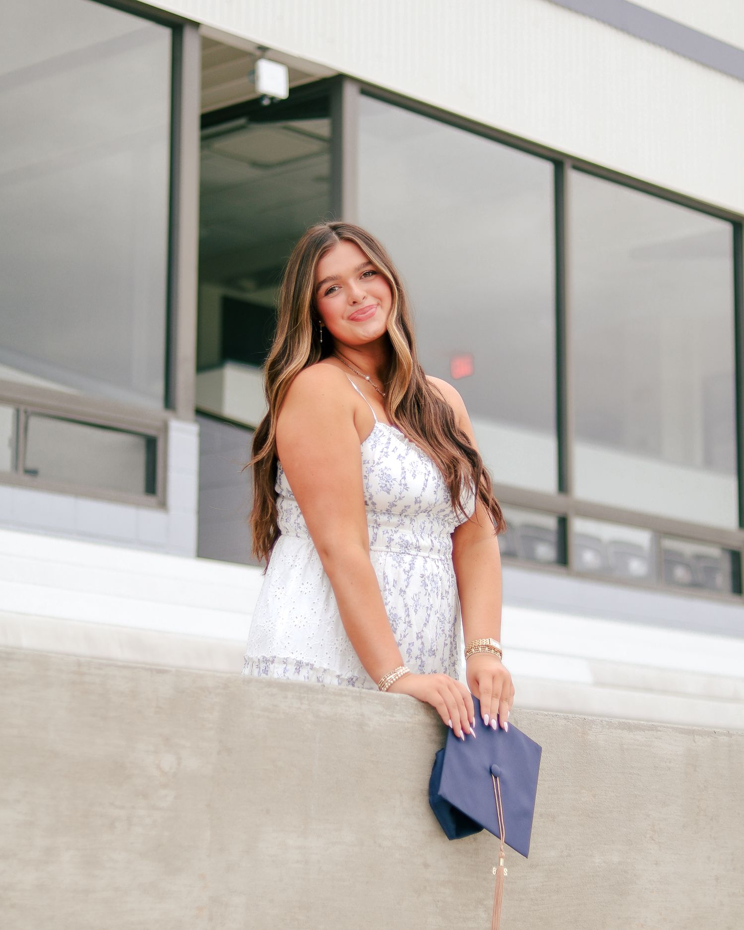 Smiling young woman with long wavy hair in a white sundress leans on a concrete ledge holding a navy graduation cap.