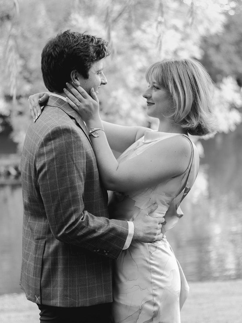 Black and white engagement photo of a smiling couple embracing by a tranquil lake, framed by soft willow branches.