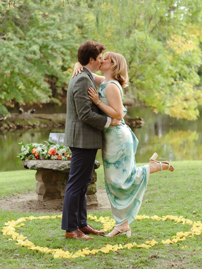 A couple shares a romantic kiss inside a circle of yellow flower petals near a pond, with a bouquet on a stone pedestal.