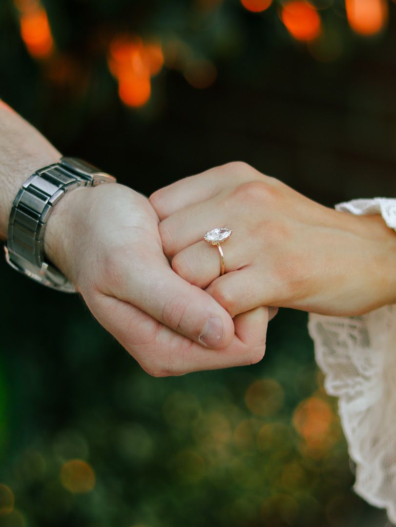 Couple holding hands showing a marquise diamond engagement ring on a rose gold band, with a silver watch and warm bokeh lights behind.