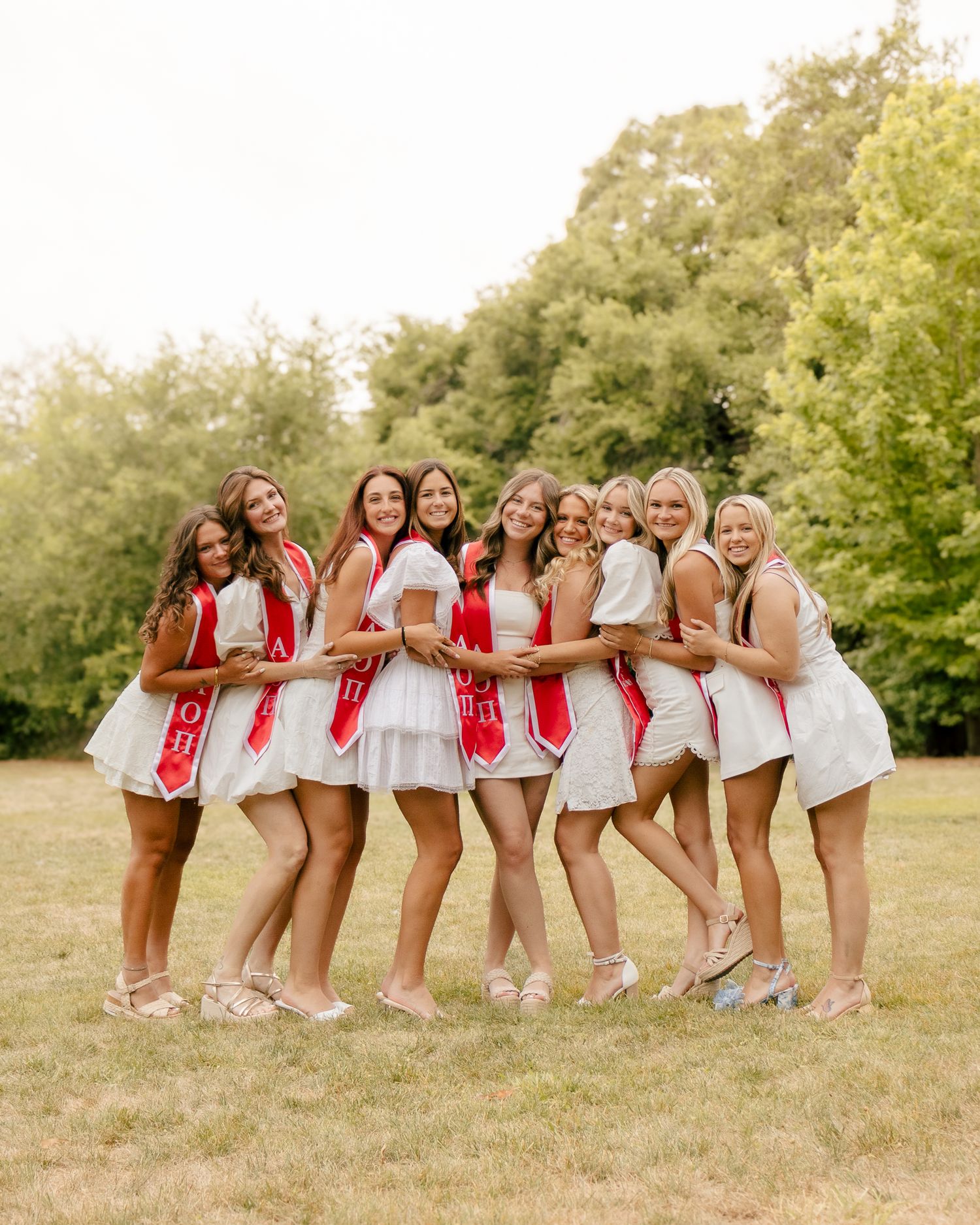 Group of young women in white dresses and red sashes pose arm in arm in a sunlit grassy park with tall green trees behind them.
