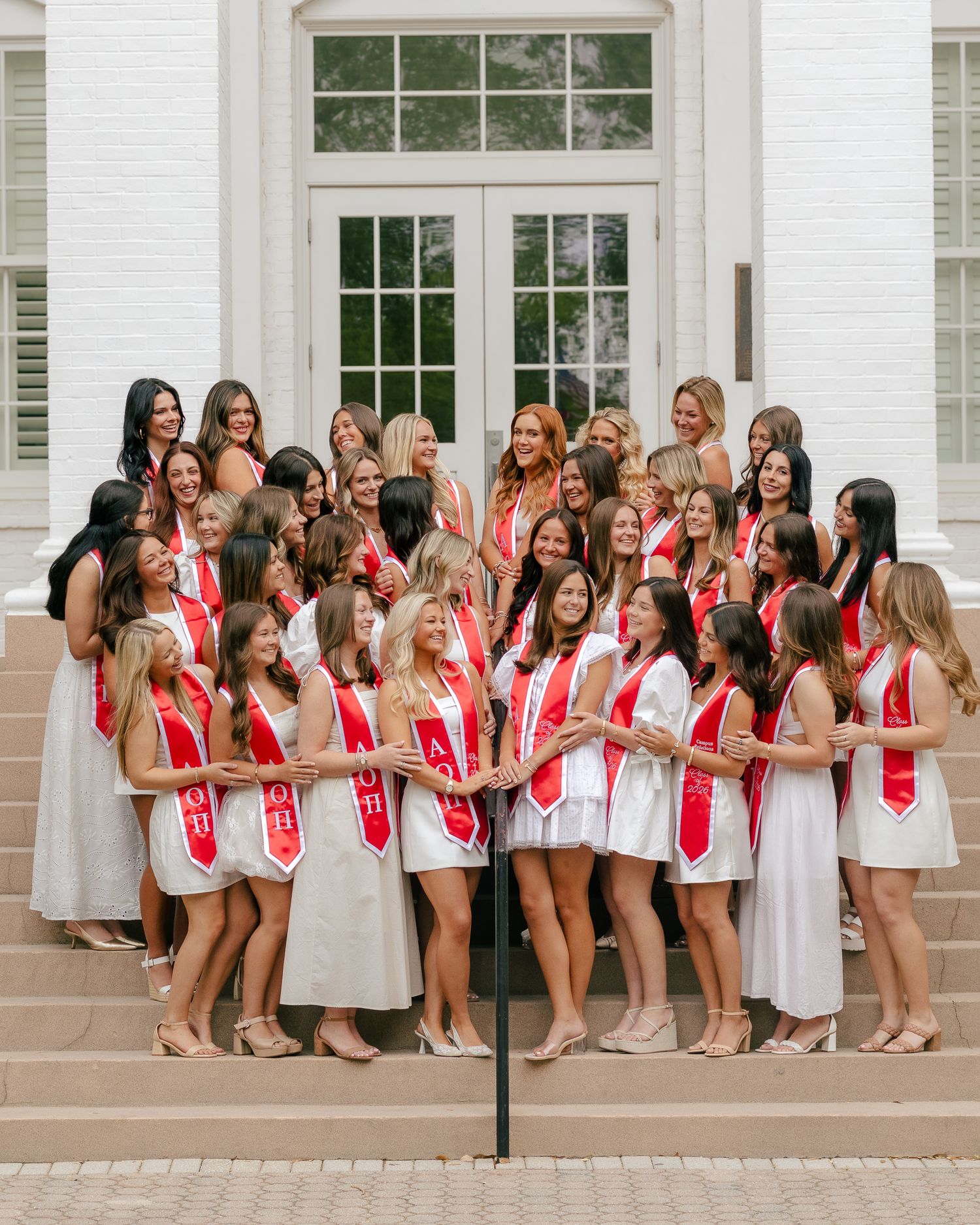 Group of smiling women in white dresses and red sashes posing together on brick steps in front of a white building with French doors.