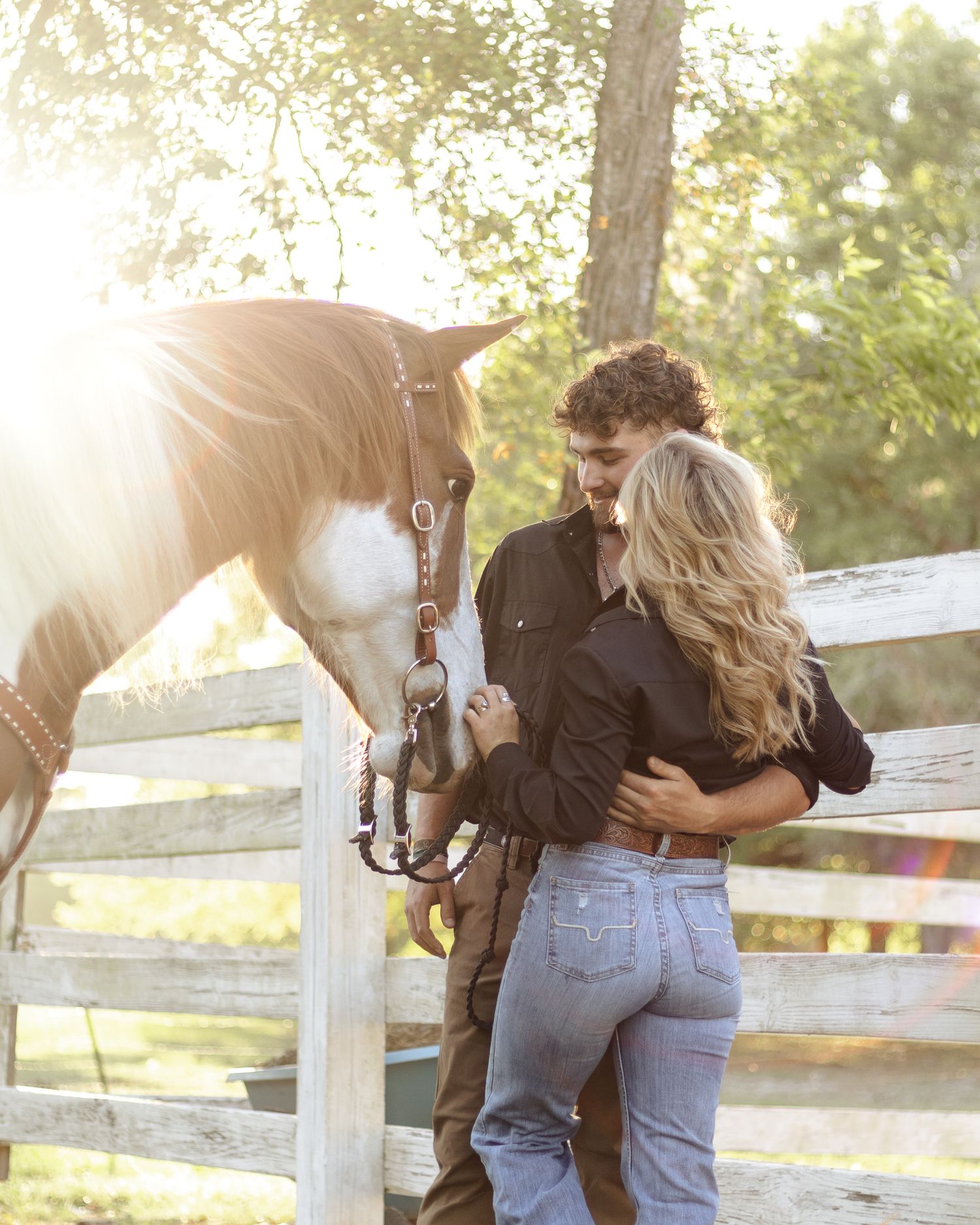 A couple embraces beside a white wooden fence while a brown and white horse in a bridle leans in close, lit by warm golden sunlight.